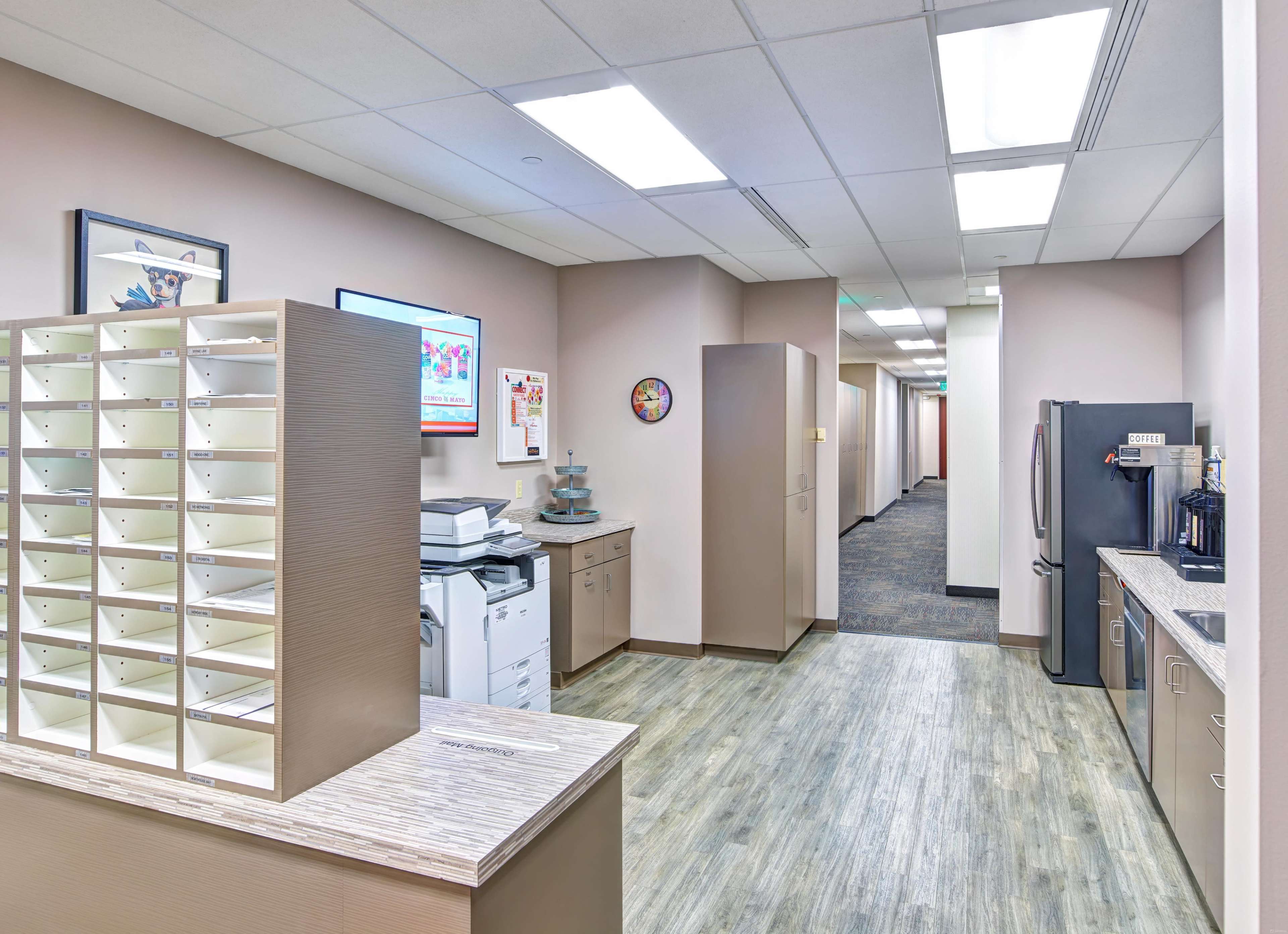 The image shows a brightly lit office hallway featuring a mail sorter, copiers, and a kitchenette area.
