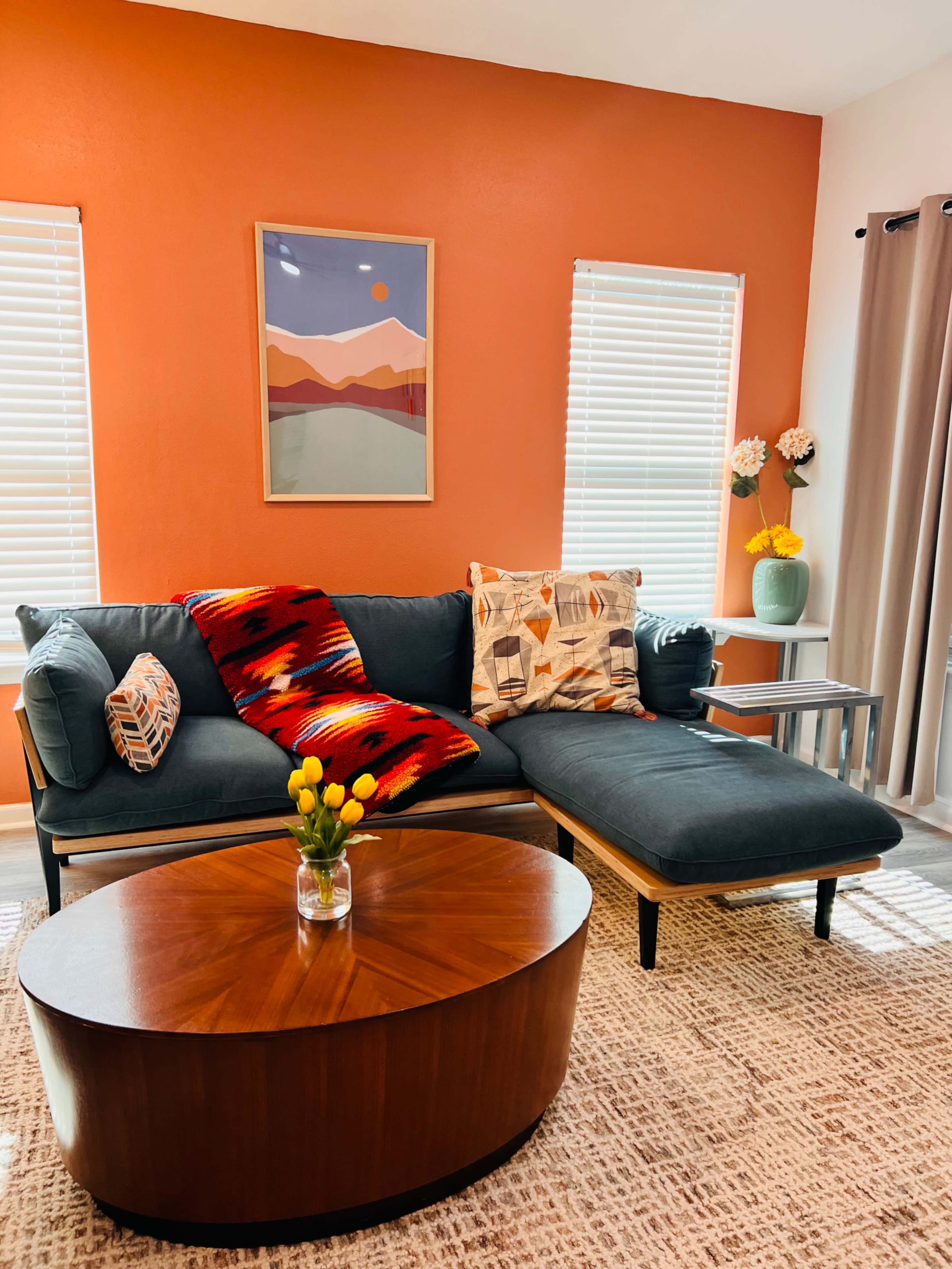 A modern living room features a sectional sofa with colorful throw pillows, a round wooden coffee table, and a vase of flowers on a side table against an orange wall.