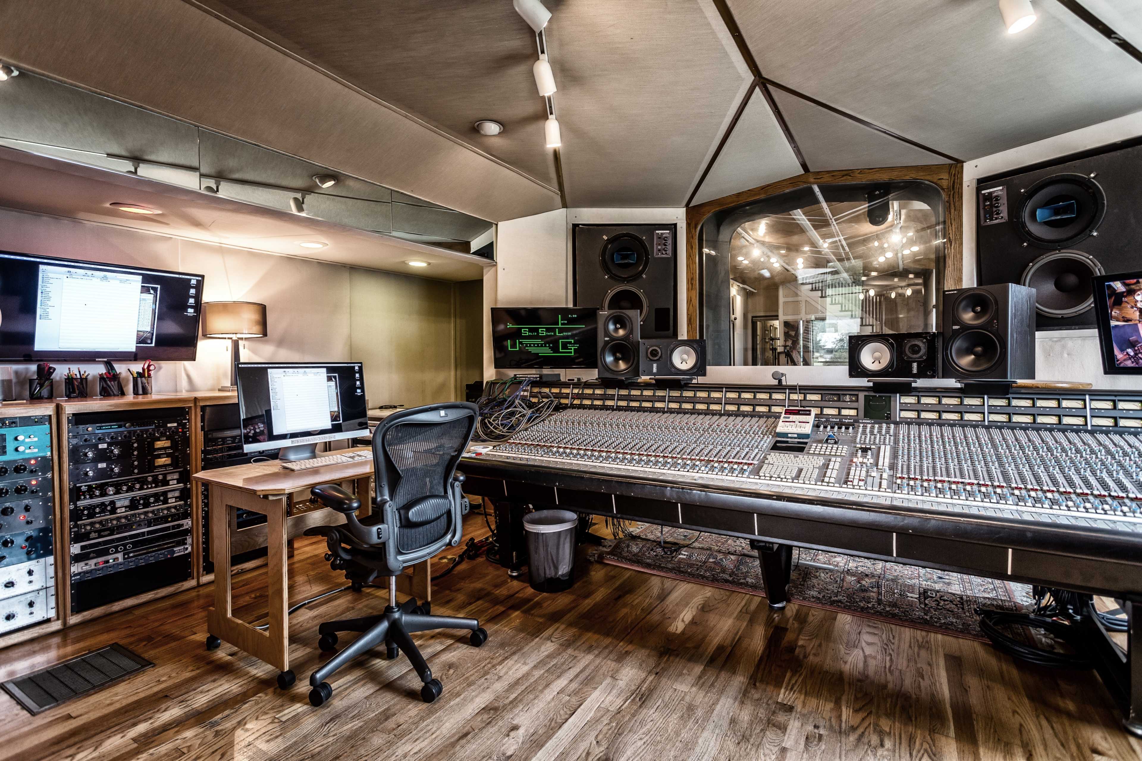 A modern recording studio featuring a large mixing console, computer monitors, and professional audio equipment arranged in a spacious room with wooden flooring.