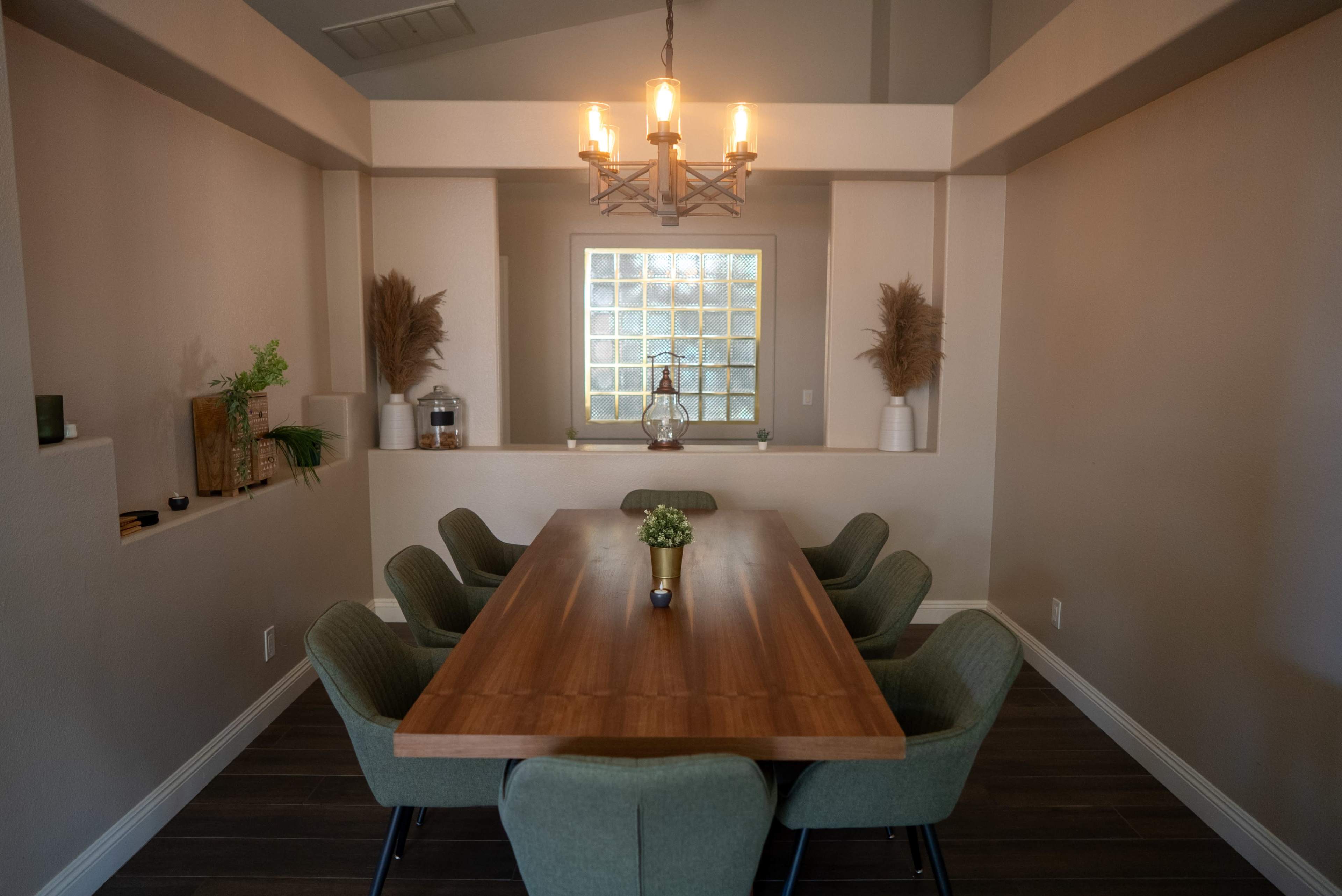 A spacious dining room features a long wooden table surrounded by eight green chairs, with a chandelier above and decorative plants along the walls.