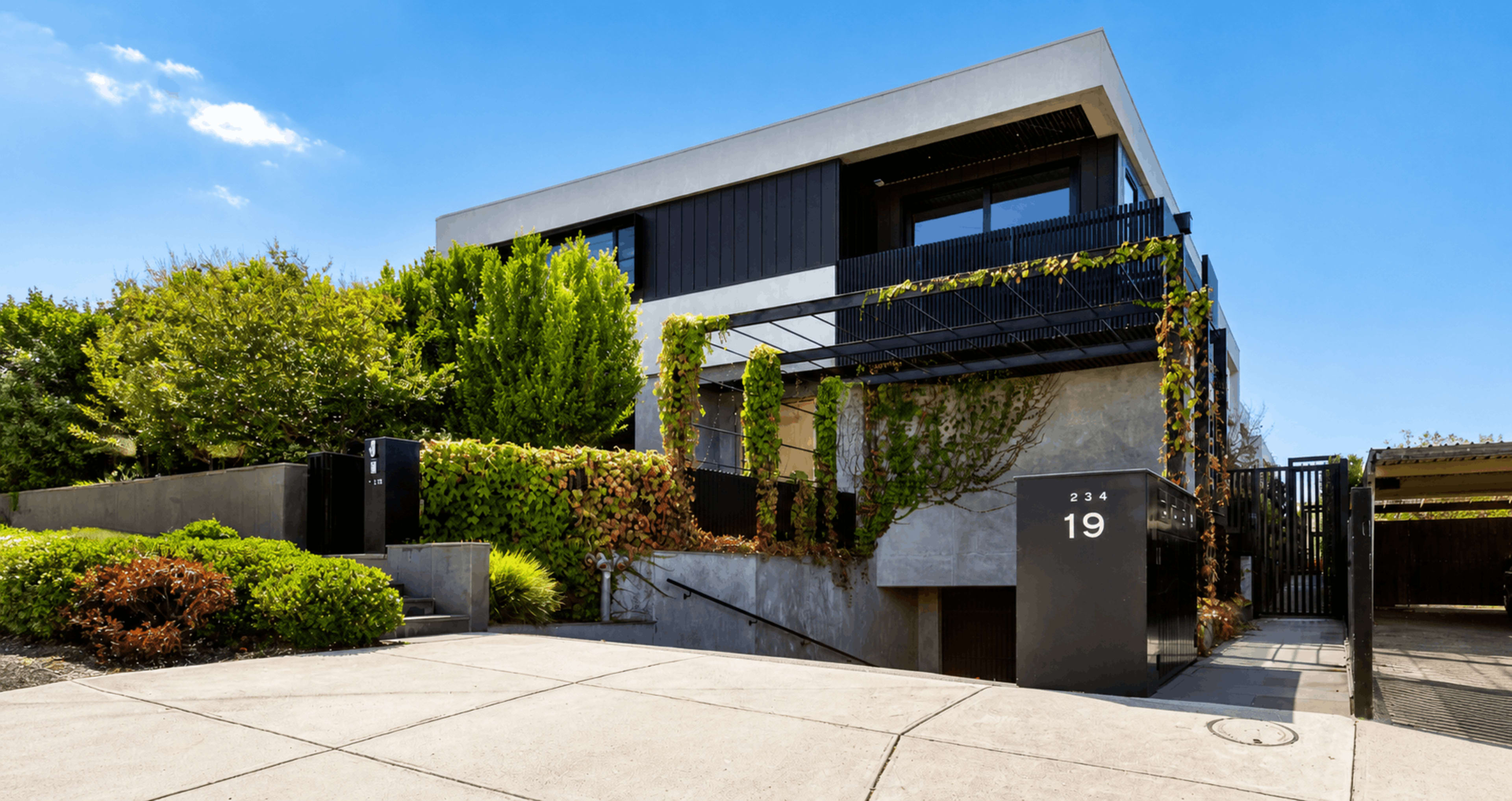 The image shows a modern two-story house with a mix of dark and light exterior materials, surrounded by lush greenery and a driveway leading to a prominent address sign.
