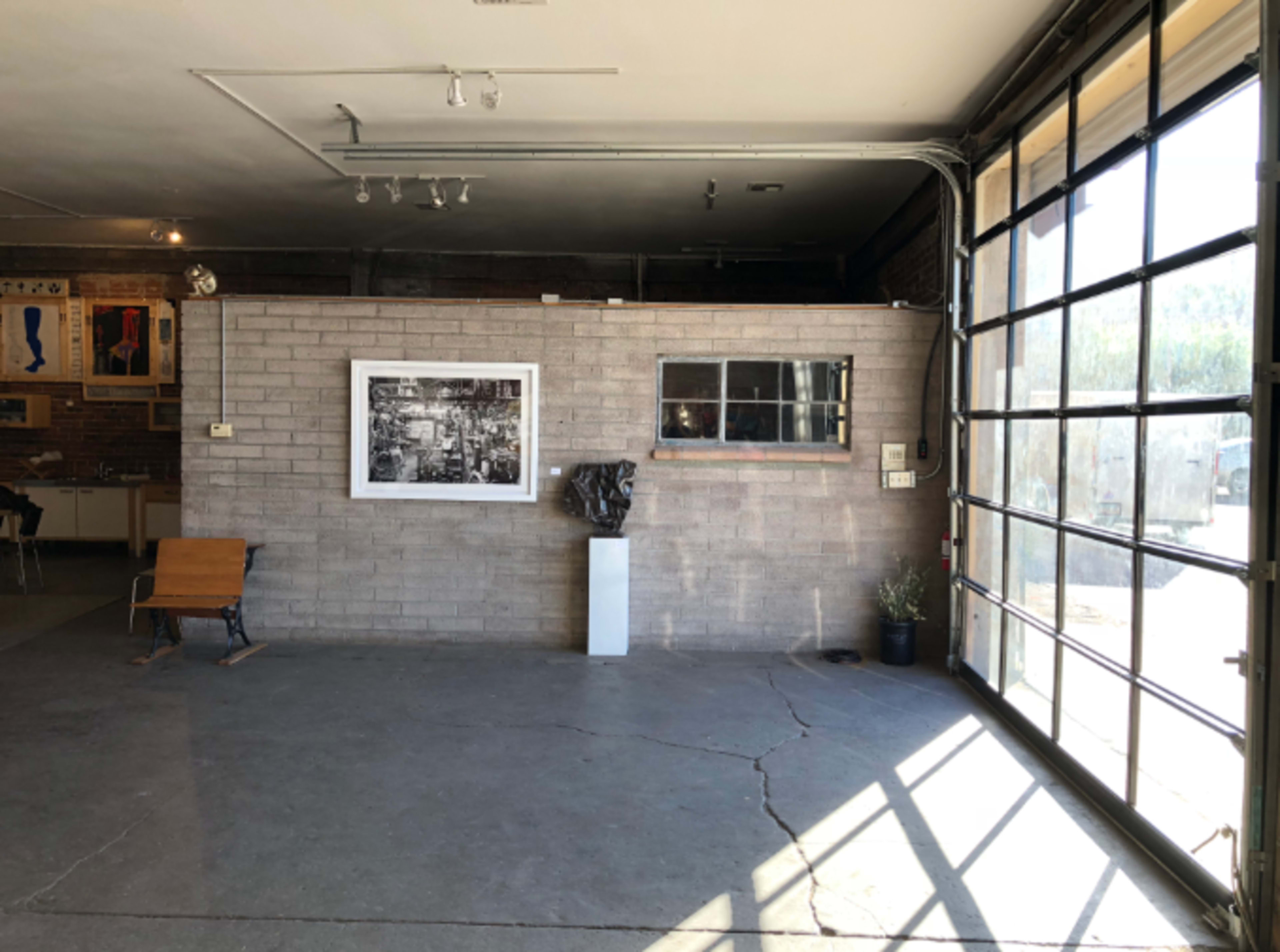 A spacious room with a concrete floor, featuring a large windowed garage door, a framed black-and-white photograph on a gray wall, and a sculpture displayed on a pedestal.