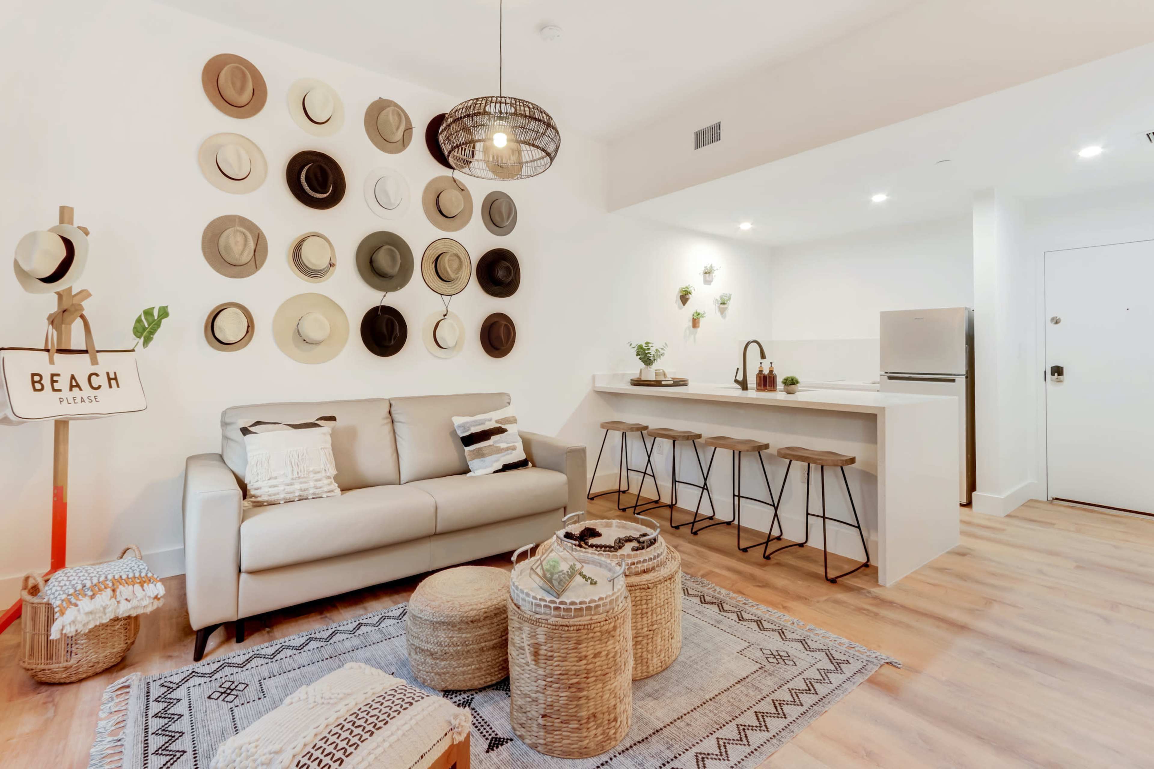 A modern living area features a light-colored sofa, a wall adorned with various hats, a kitchen counter with stools, and decorative elements like potted plants and a beach-themed sign.