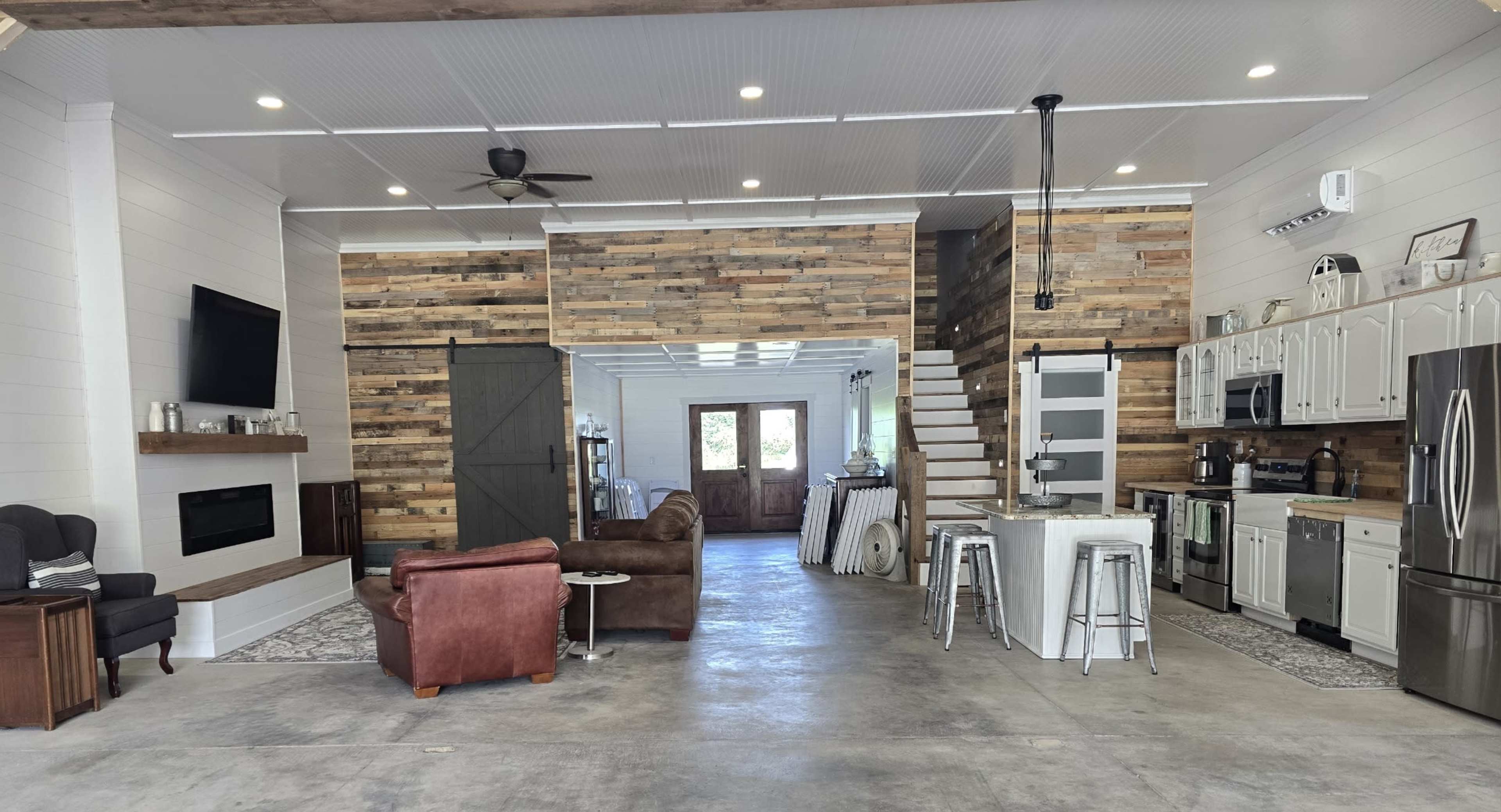 The interior of a modern open-concept home features a living area with a brown leather sofa, a kitchen with stainless steel appliances, and wooden wall accents.