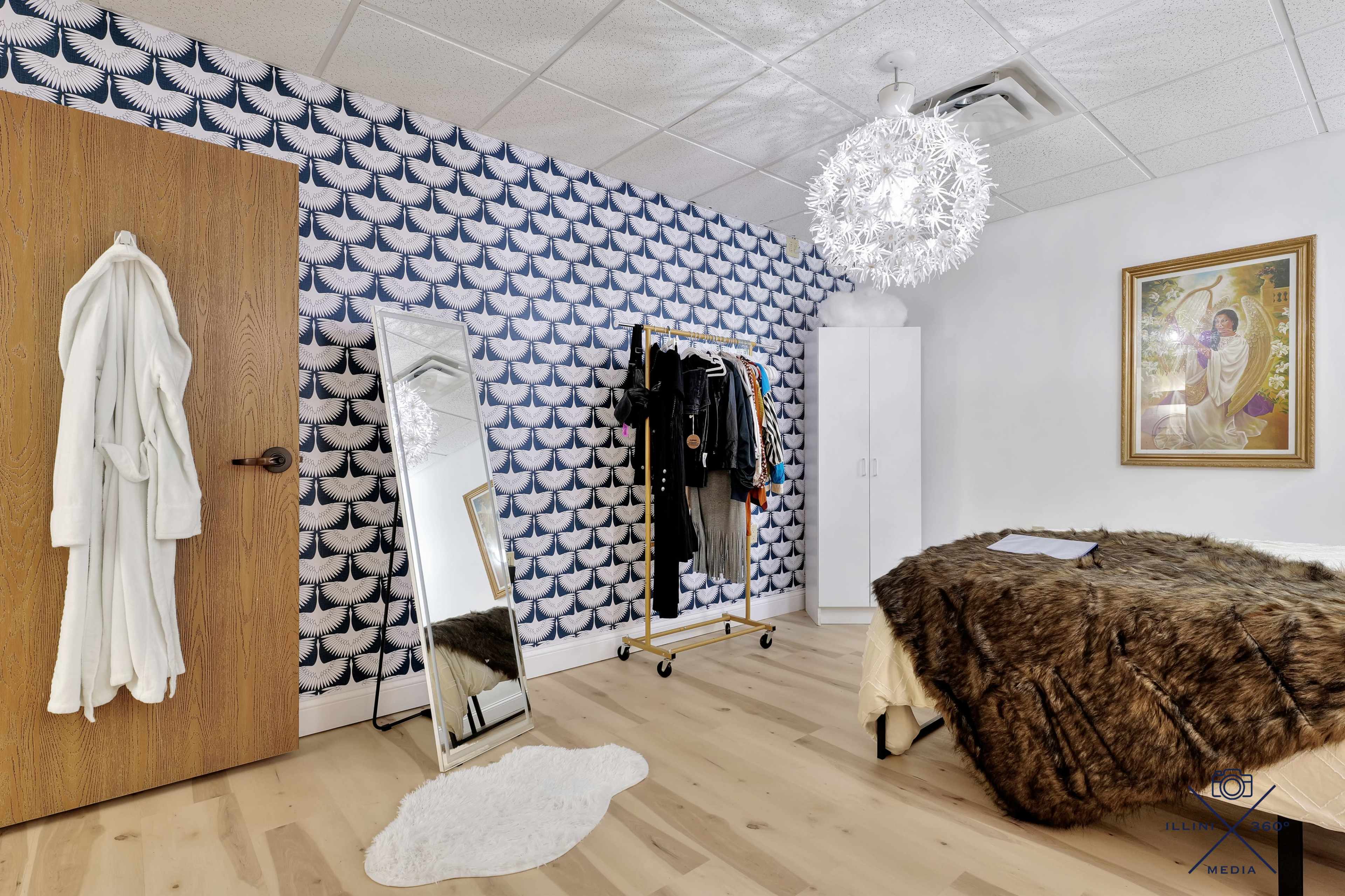 The image shows a modern bedroom featuring a patterned accent wall, a large mirror, a clothing rack, a white cabinet, and a plush bedspread.