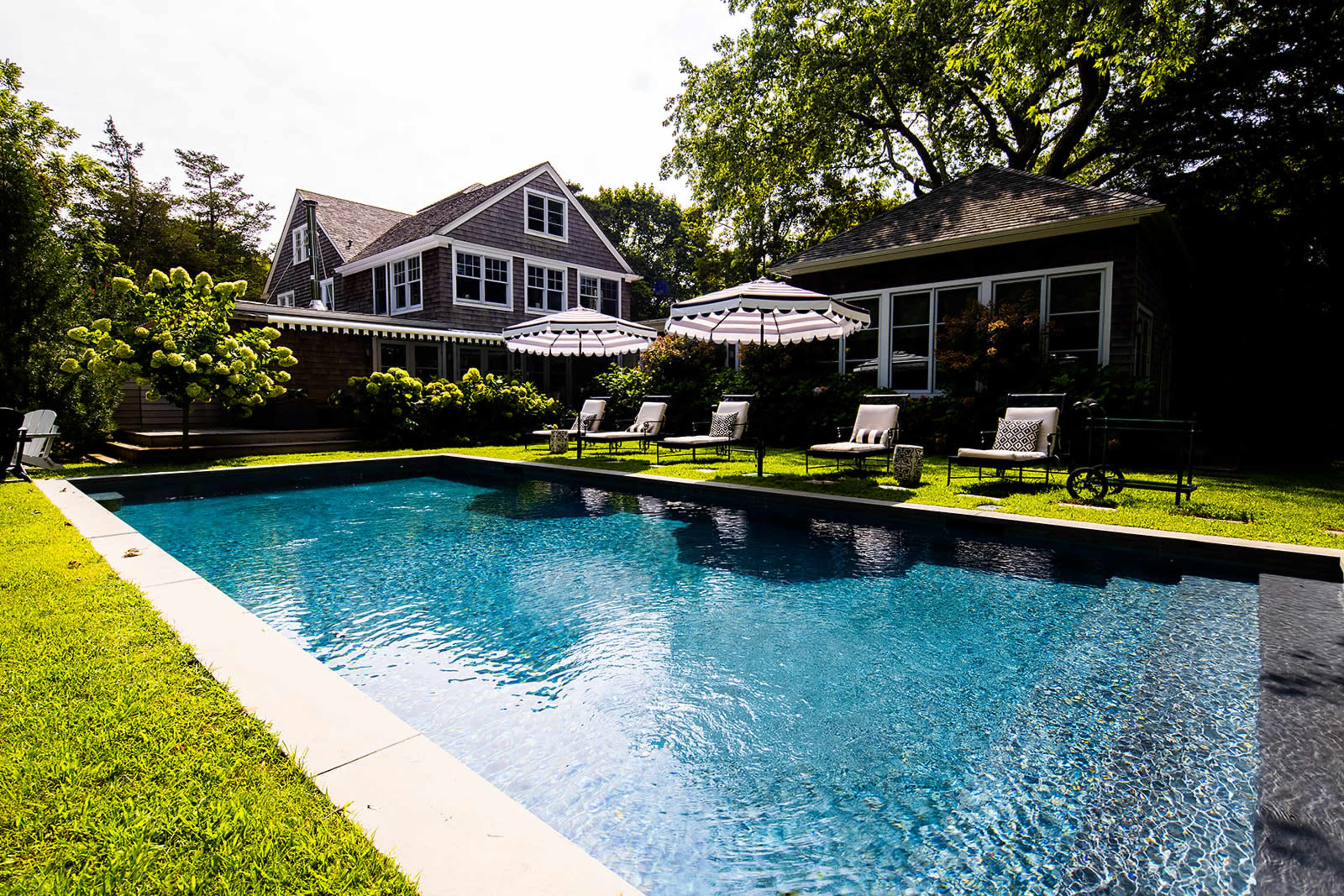 A clear swimming pool reflects the blue sky and is surrounded by lounge chairs and greenery near a house with multiple gabled roofs.