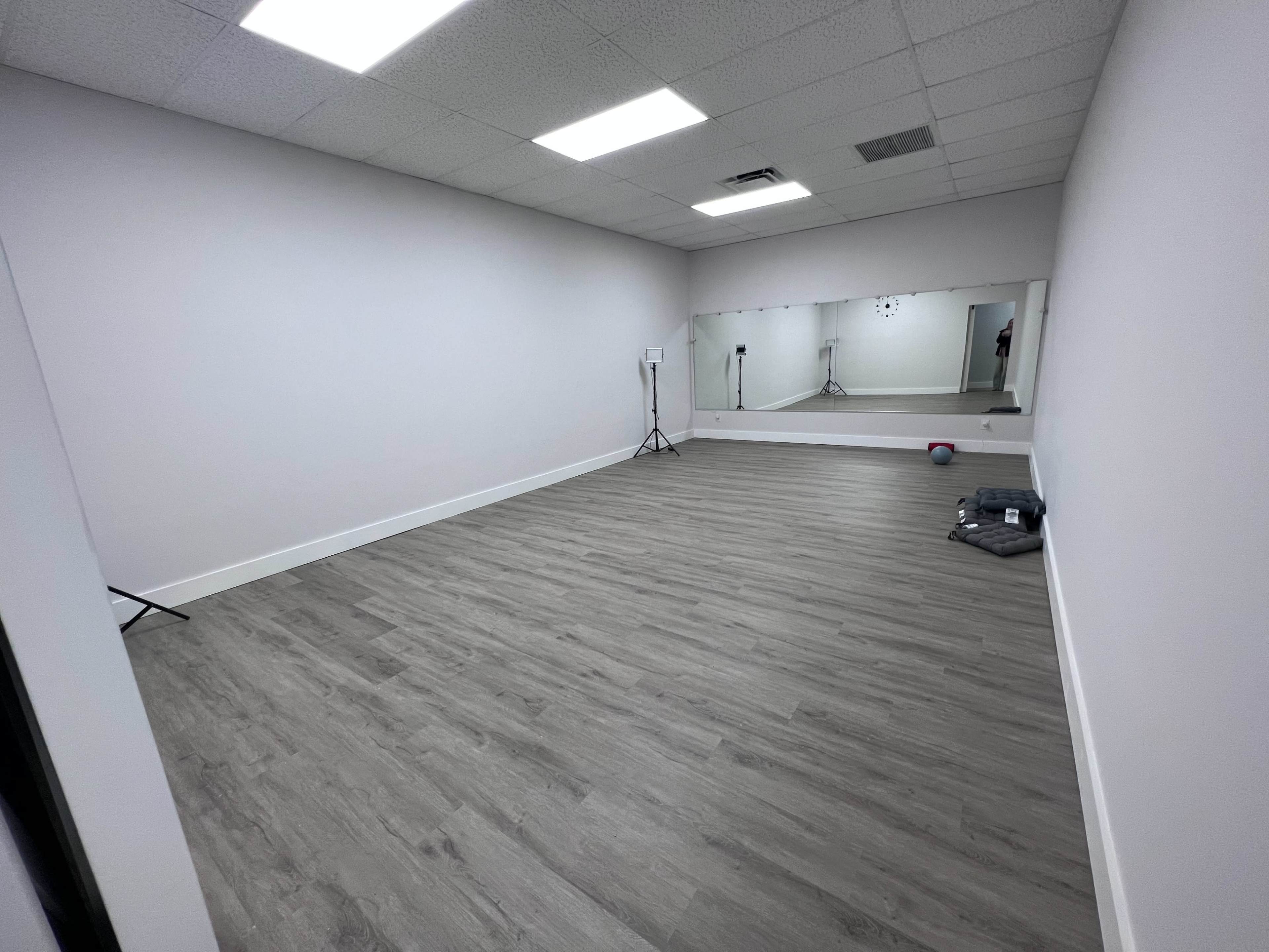 A spacious, empty room with light-colored walls, a mirrored wall, and wooden flooring, equipped with a few scattered props and lighting equipment.