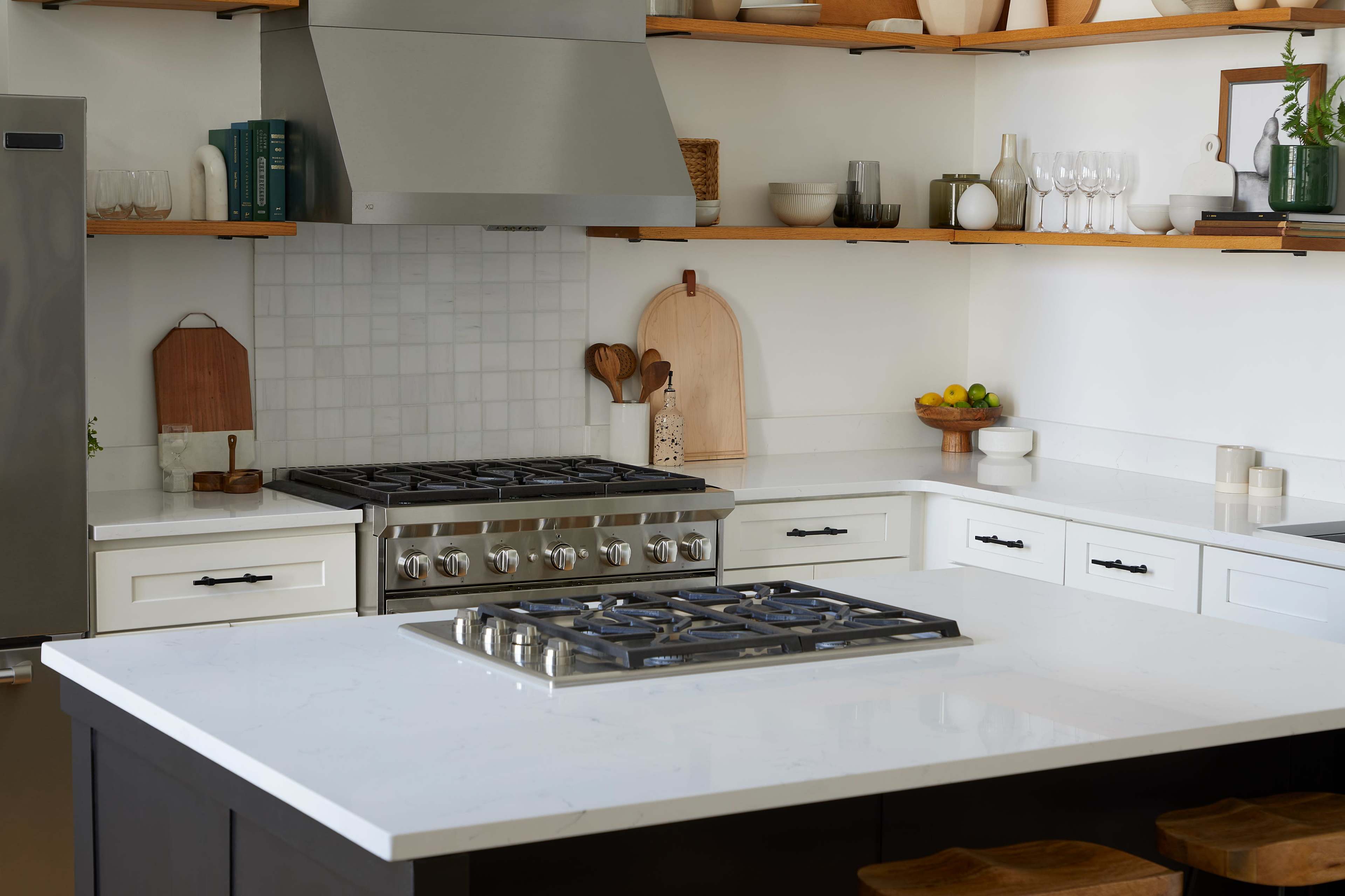 The image shows a modern kitchen with a gas stove, white countertops, wooden shelves, and various kitchen items displayed.