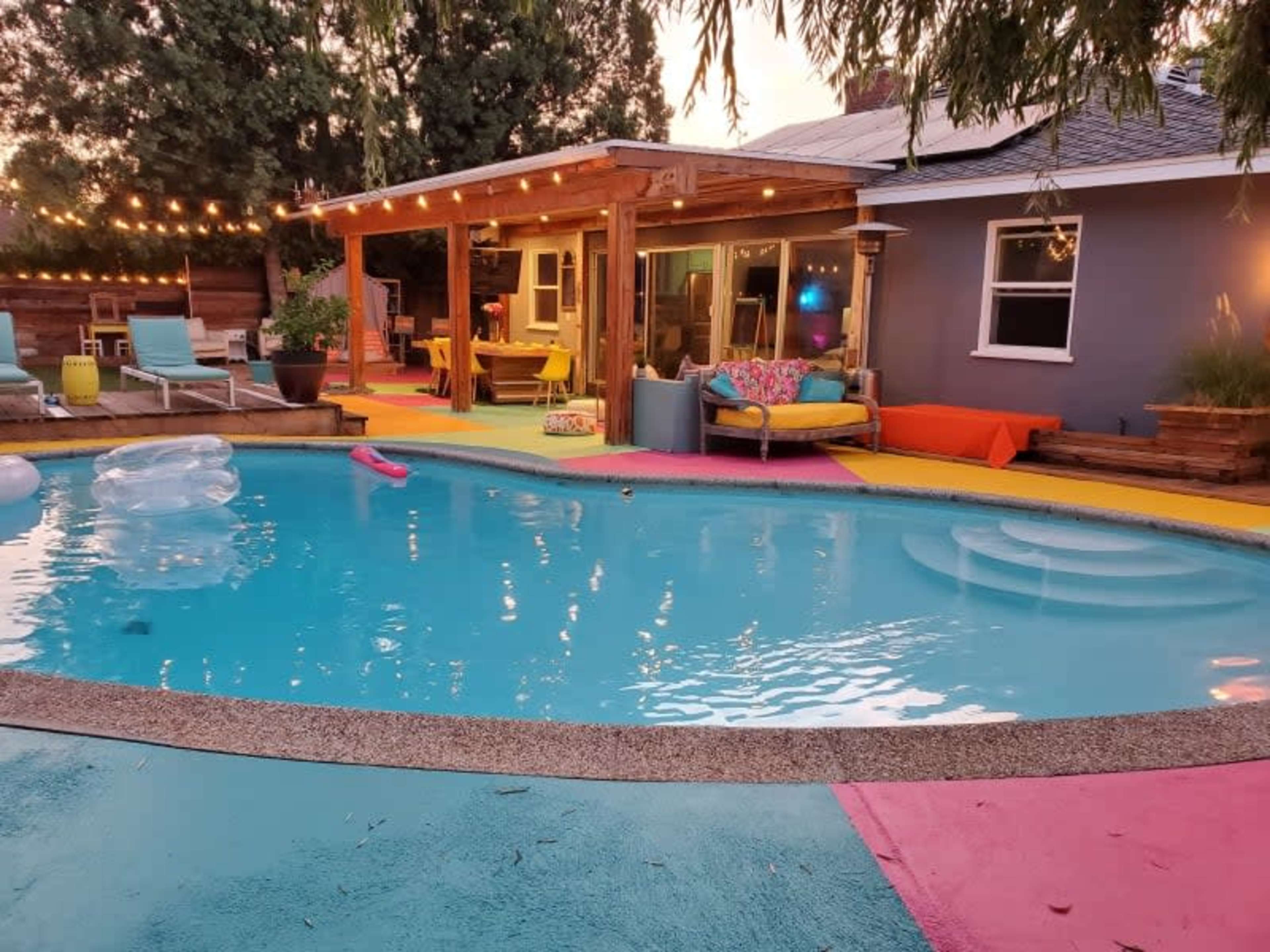The image shows a brightly colored backyard pool area with a wooden gazebo, lounge chairs, and decorative string lights.