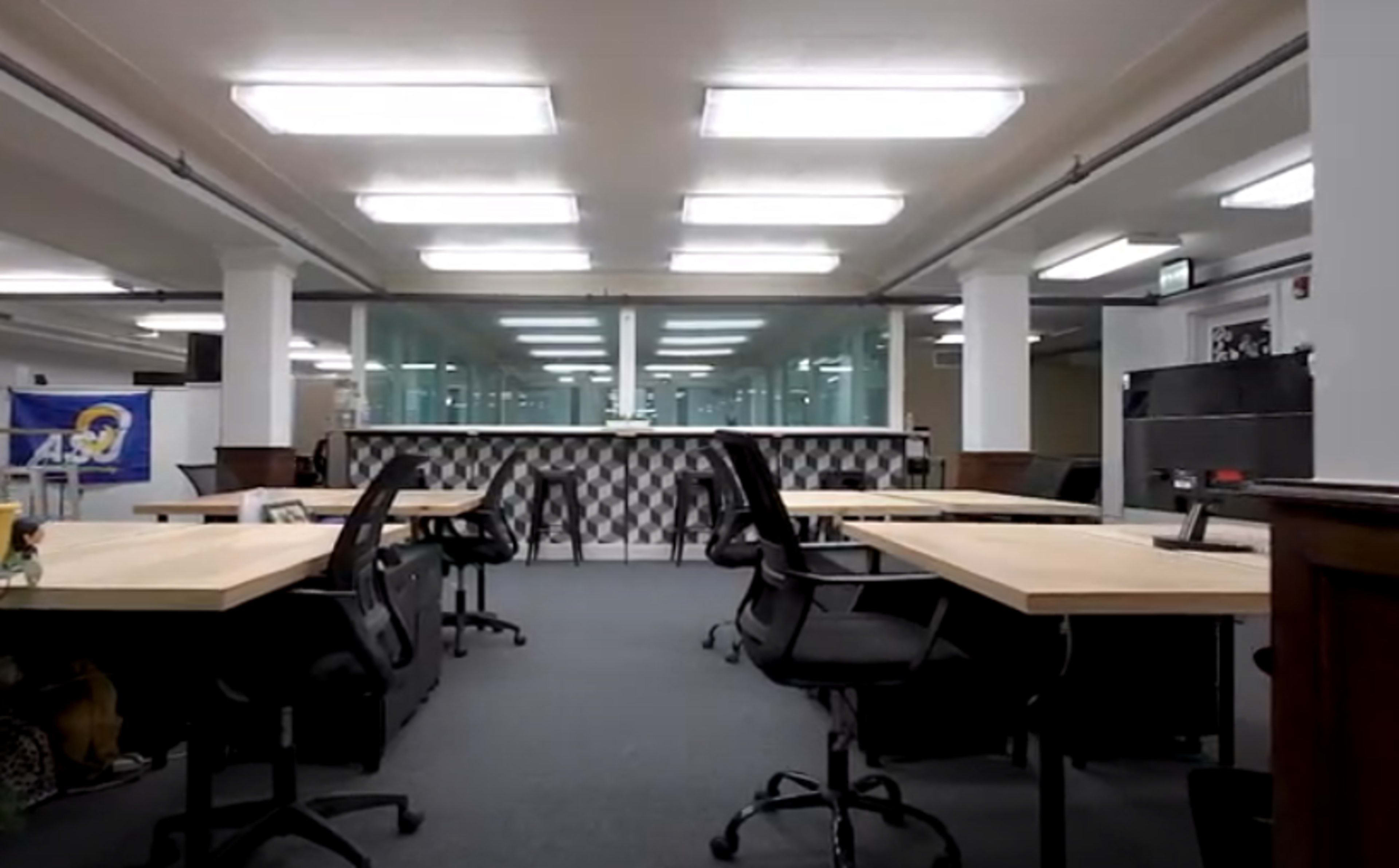 A modern office space with multiple wooden desks and ergonomic chairs arranged facing a reception area, against a backdrop of large glass windows.