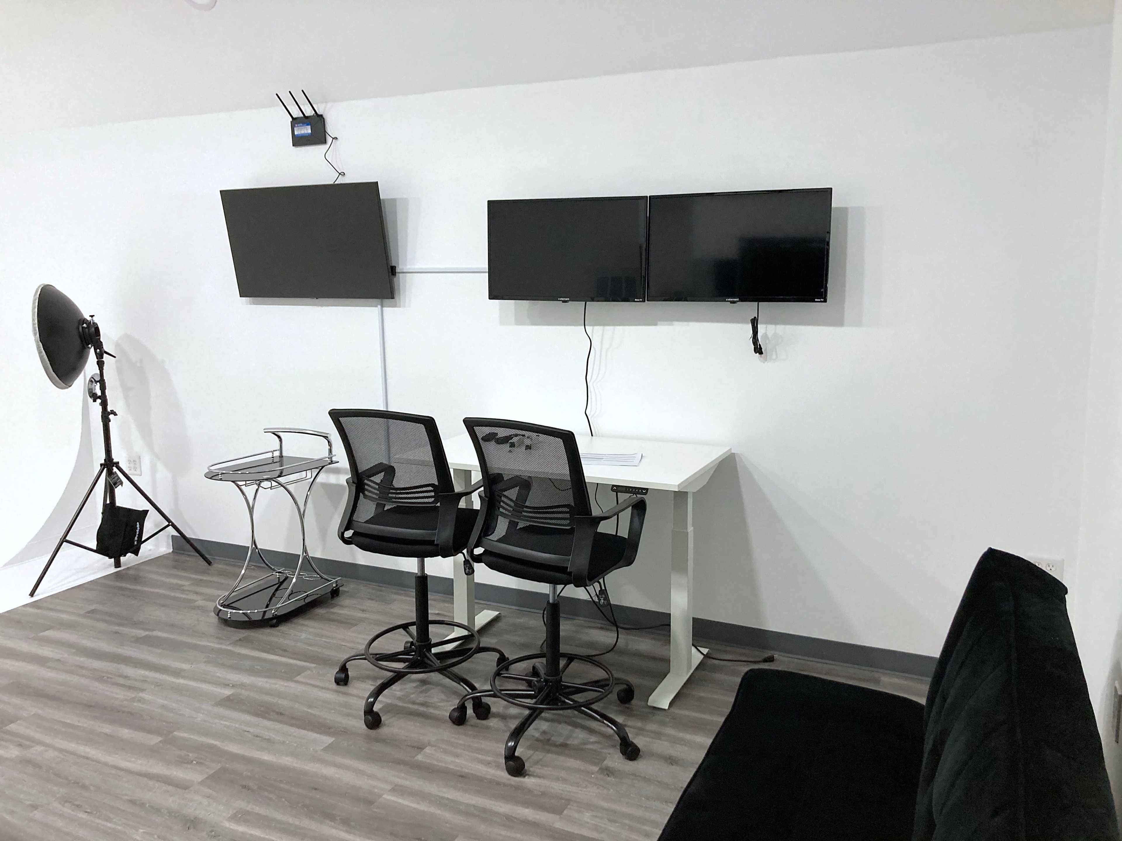 The image shows a minimalistic workspace with two black chairs, a white desk, two mounted monitors on the wall, and a softbox light.