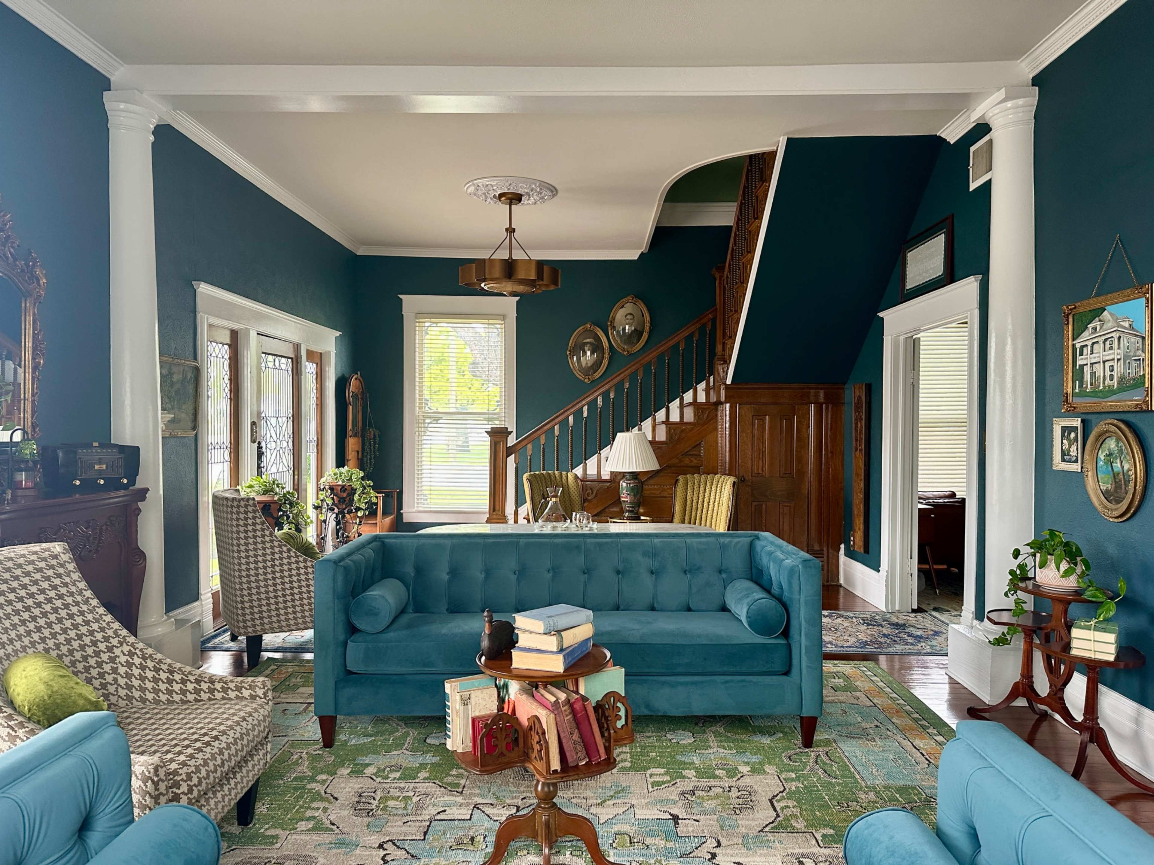 The image shows a well-furnished living room featuring a teal couch, vintage decor, a staircase in the background, and large windows allowing natural light.