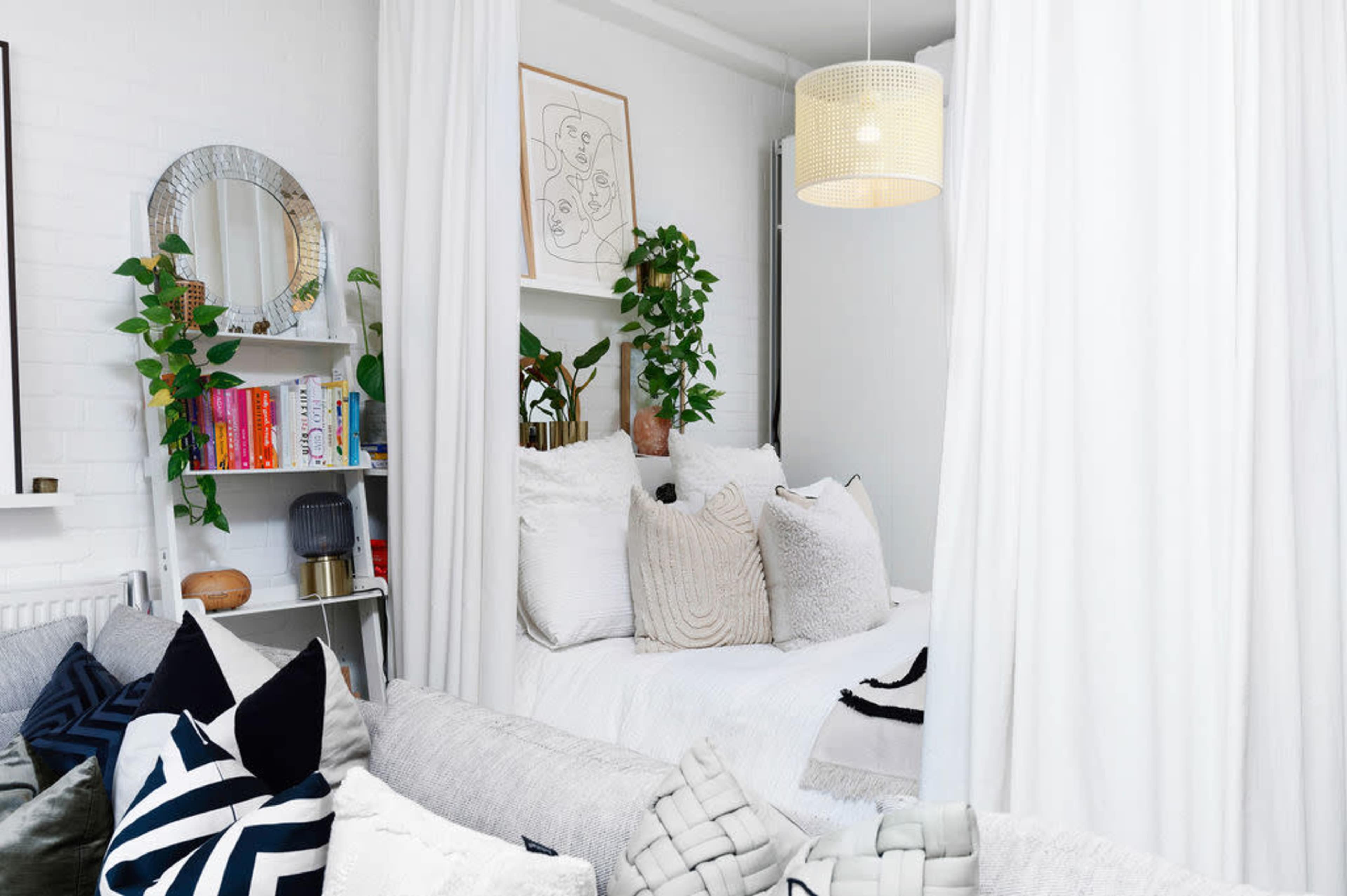 A cozy bedroom area is separated by white curtains, featuring a bed with multiple pillows and surrounded by plants, bookshelves, and a mirror.