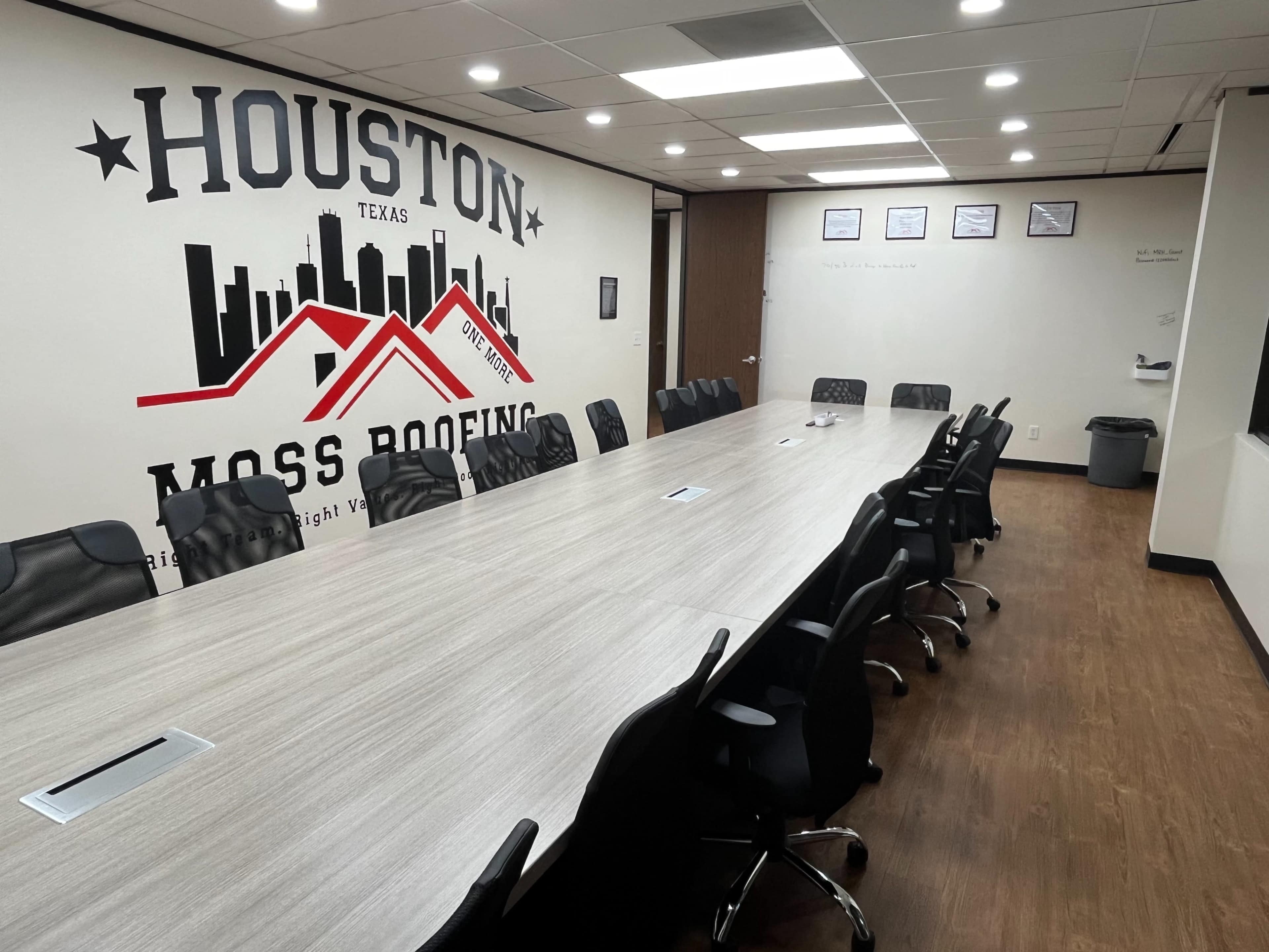 The image shows a modern conference room with a long, rectangular table surrounded by black chairs and a wall featuring a mural of the Houston skyline and company branding.