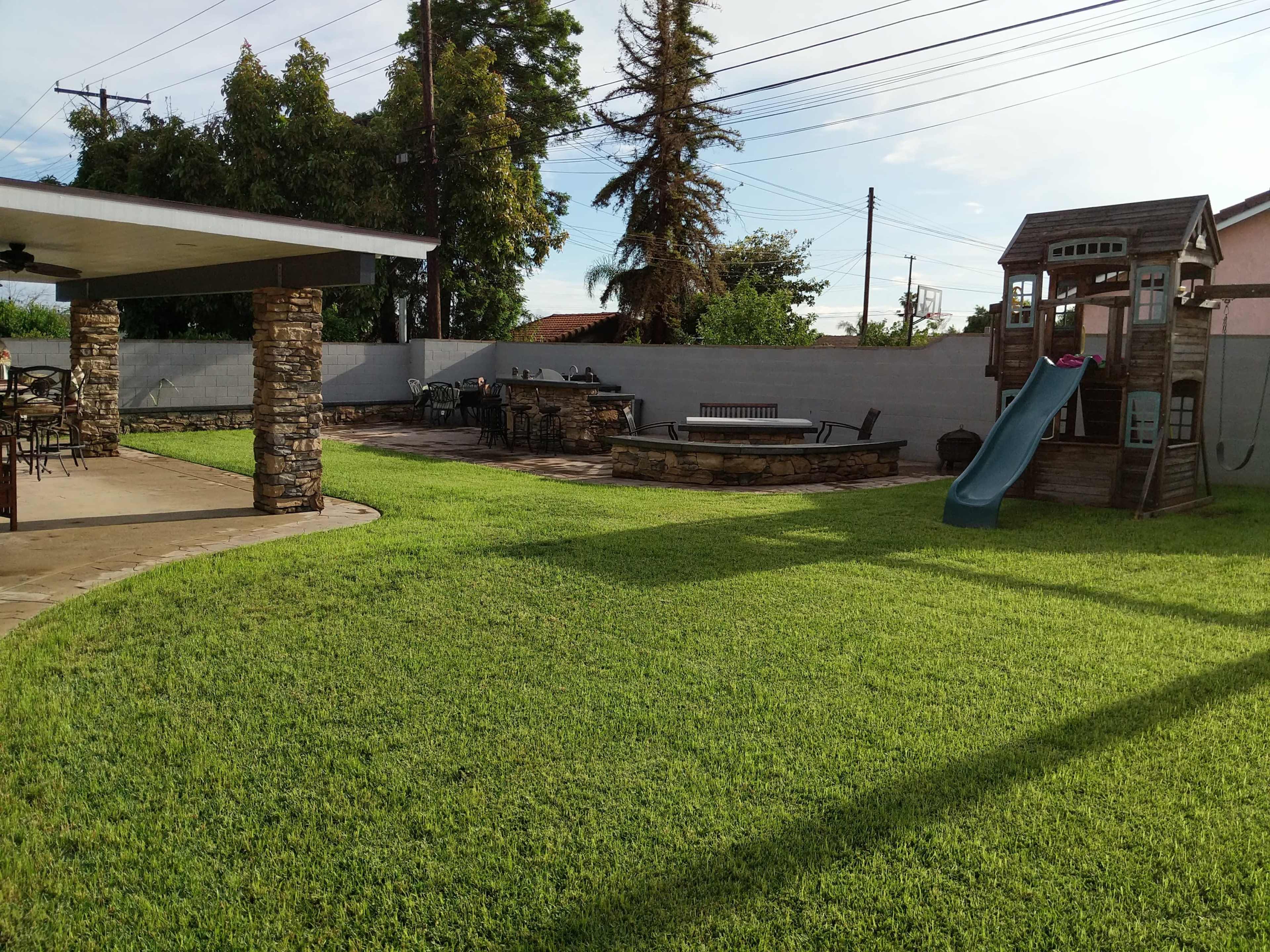 A backyard with a well-maintained lawn, a stone patio area with seating, a picnic table, and a playset with a slide.