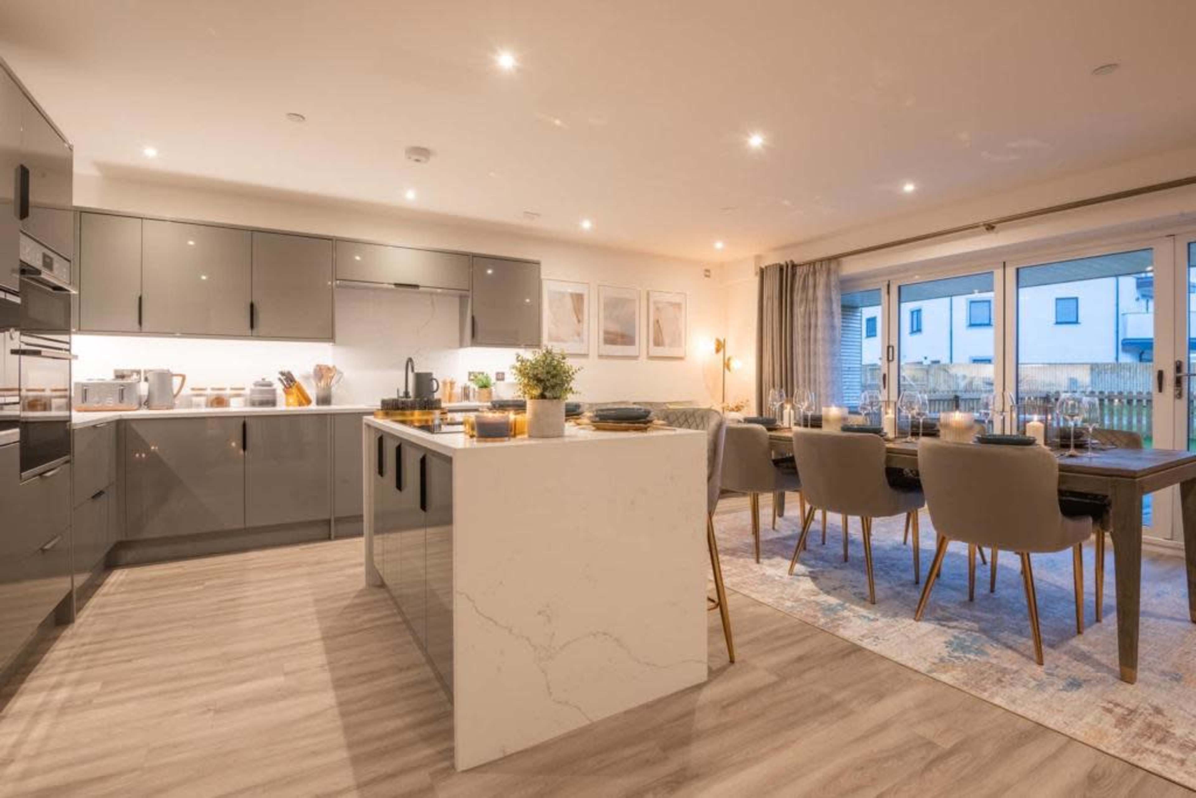A modern kitchen and dining area are shown, featuring sleek gray cabinetry, a central island with a marble countertop, and a dining table set for several guests.