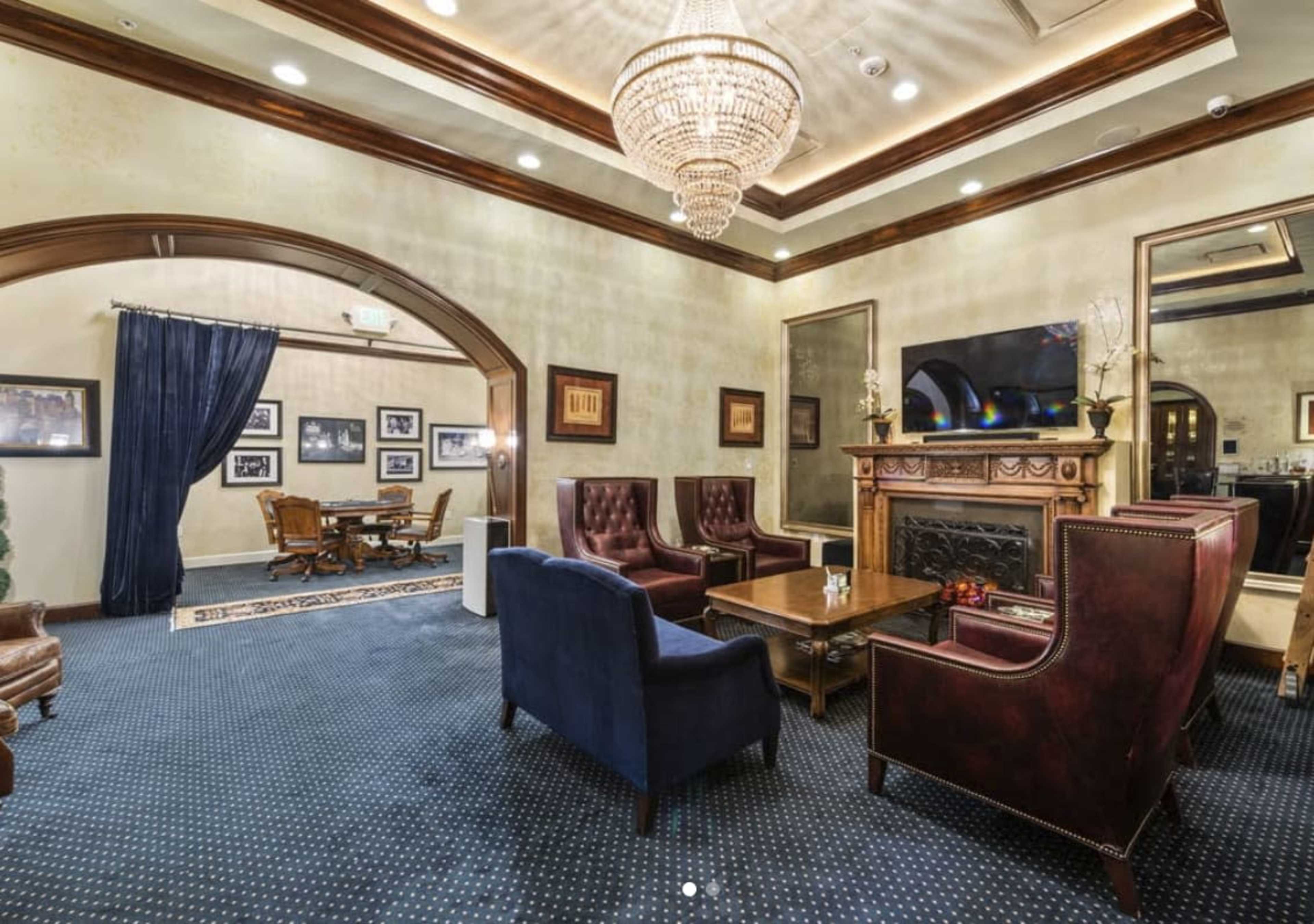 The image shows a luxurious lounge area featuring dark wooden furnishings, plush seating, and a fireplace with a chandelier overhead.