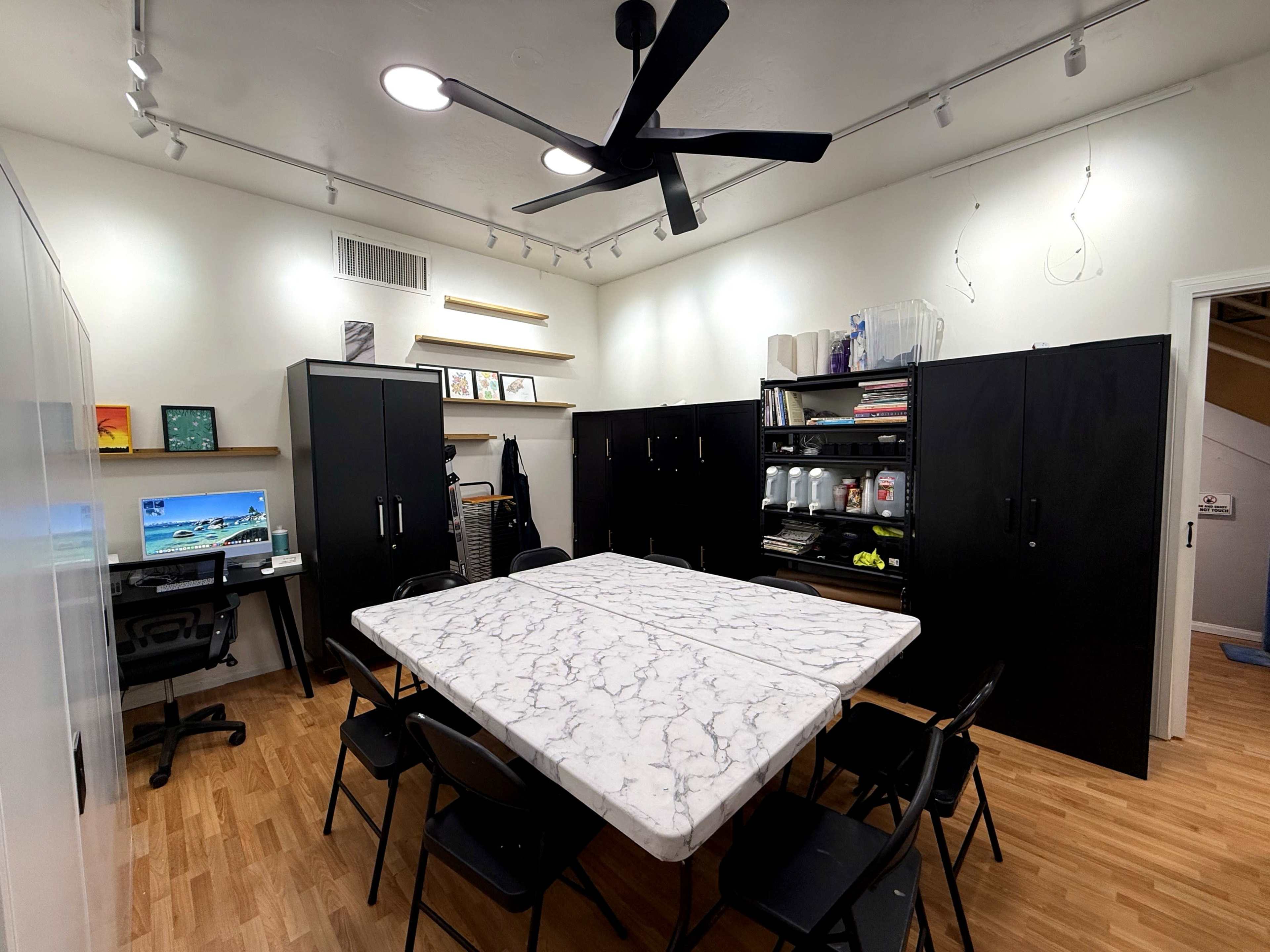 The image shows a modern office space featuring a large marble table surrounded by black folding chairs, with black cabinets lining the walls and a desk with a computer in one corner.