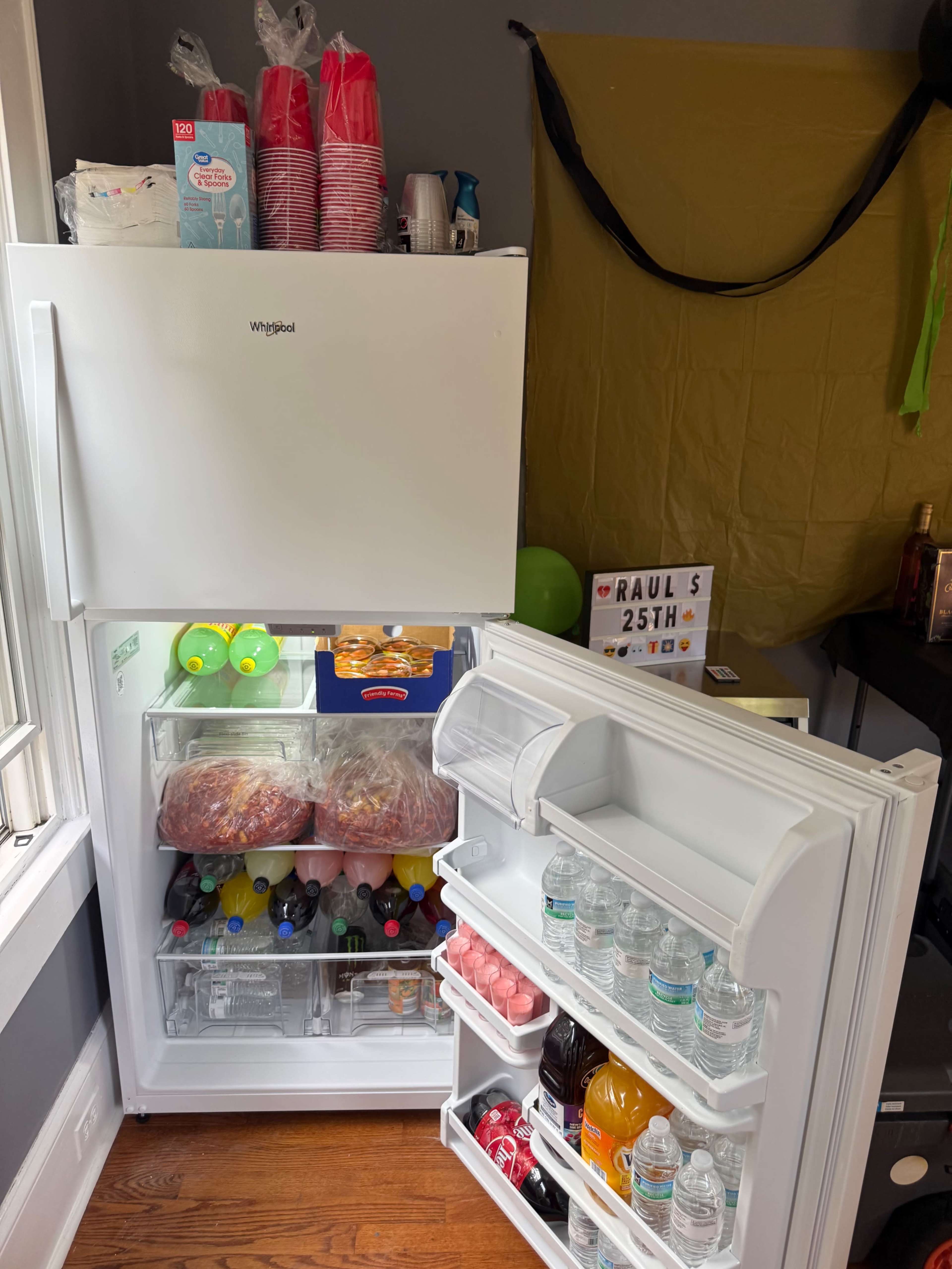 A white Whirlpool refrigerator is open, displaying a variety of drinks, including soda and bottled water, alongside food items and party supplies on top.