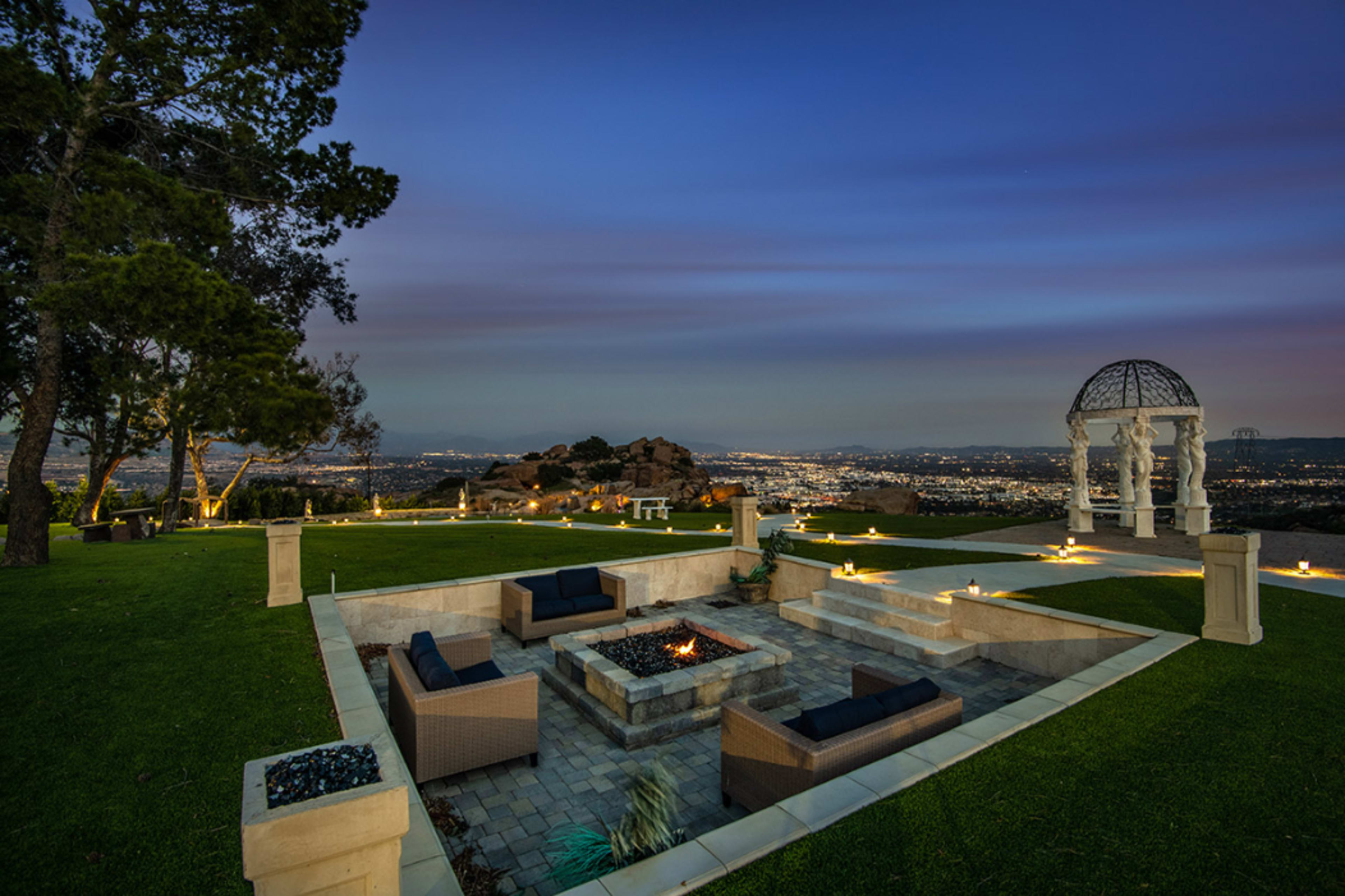 The image shows a landscaped outdoor space with seating around a fire pit, a decorative gazebo, and a panoramic view of a city below at dusk.