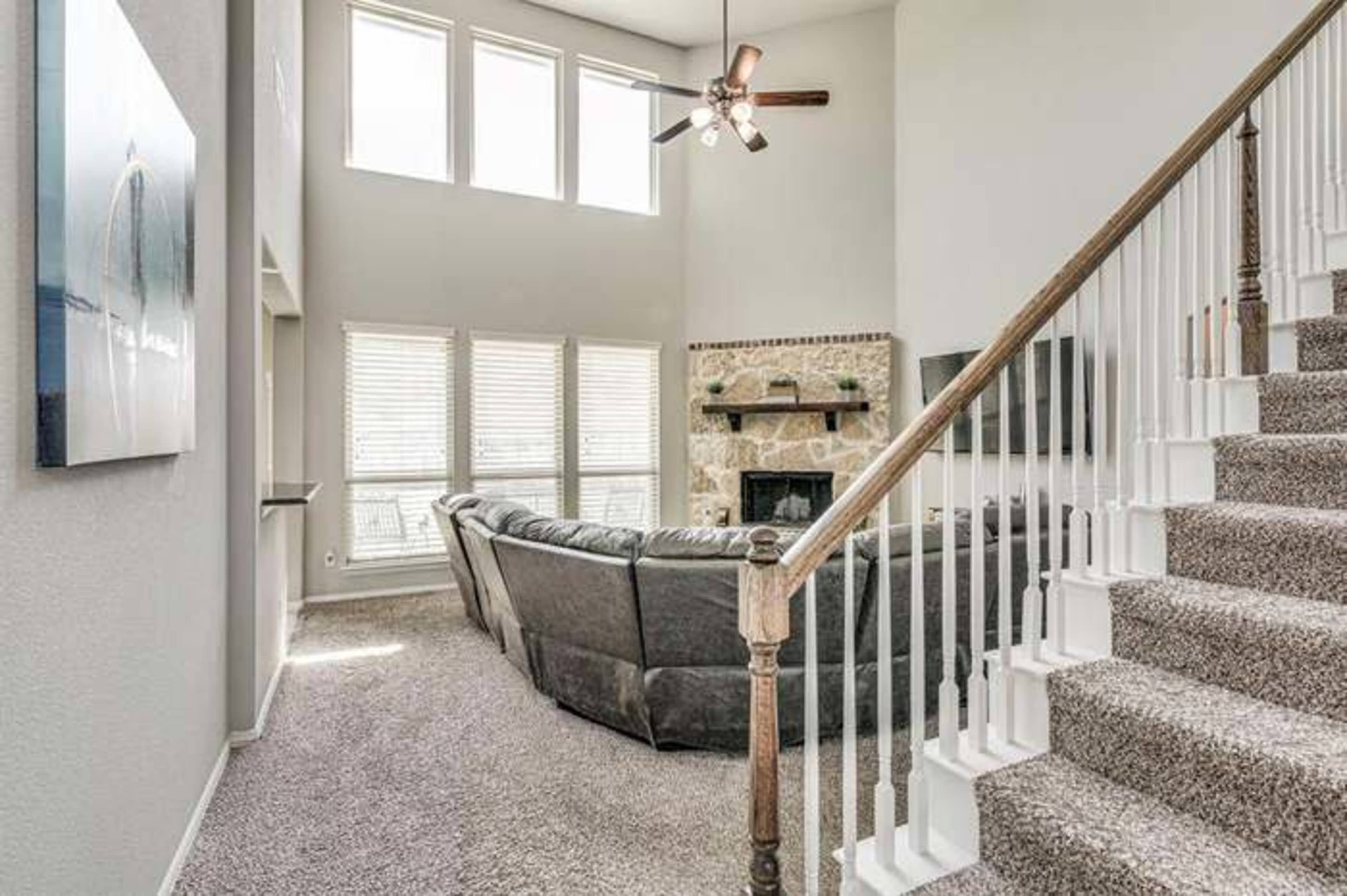 The image shows a living room with a gray sectional sofa, a stone fireplace, a flat-screen TV mounted on the wall, and a staircase leading to the upper level.