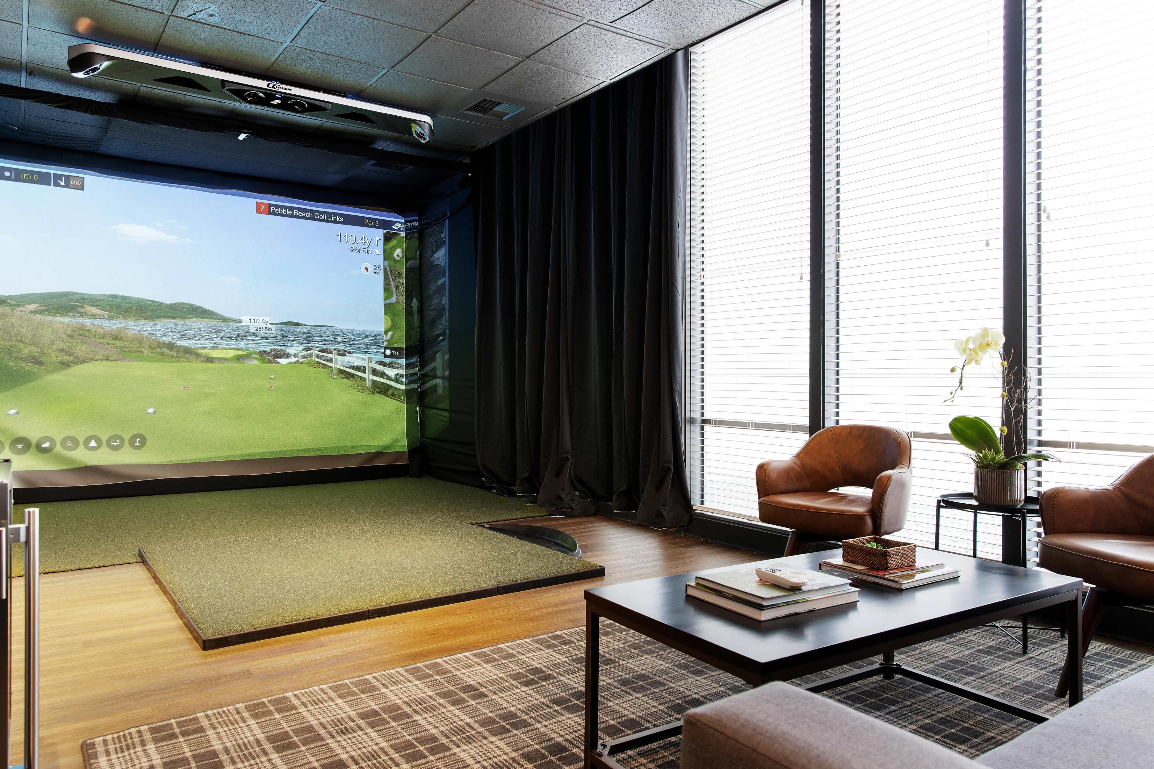 A modern indoor golf simulator setup, featuring a green turf area, a large projection screen, and two leather chairs beside a coffee table in a well-lit room.