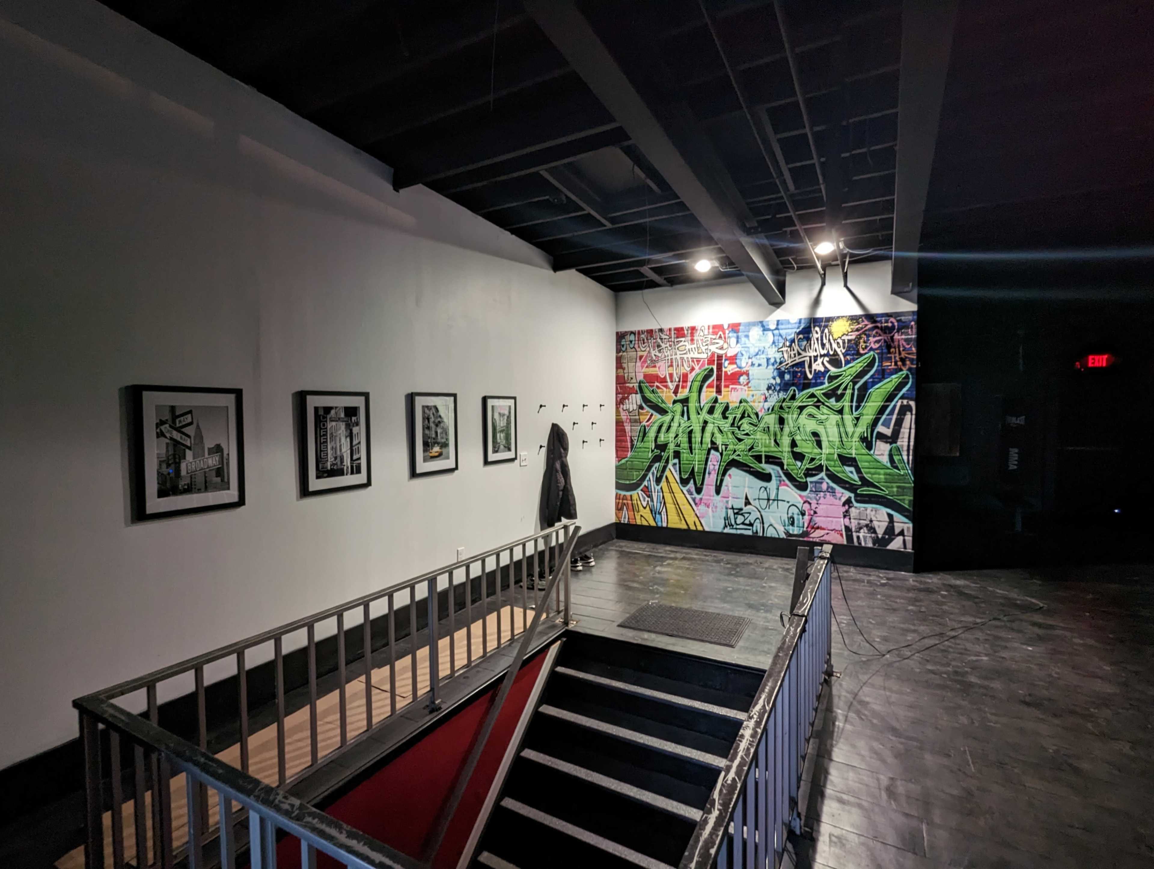An indoor space with a staircase leading down to a lower level, featuring black and white framed photographs on the wall and a colorful graffiti mural on the opposite wall.