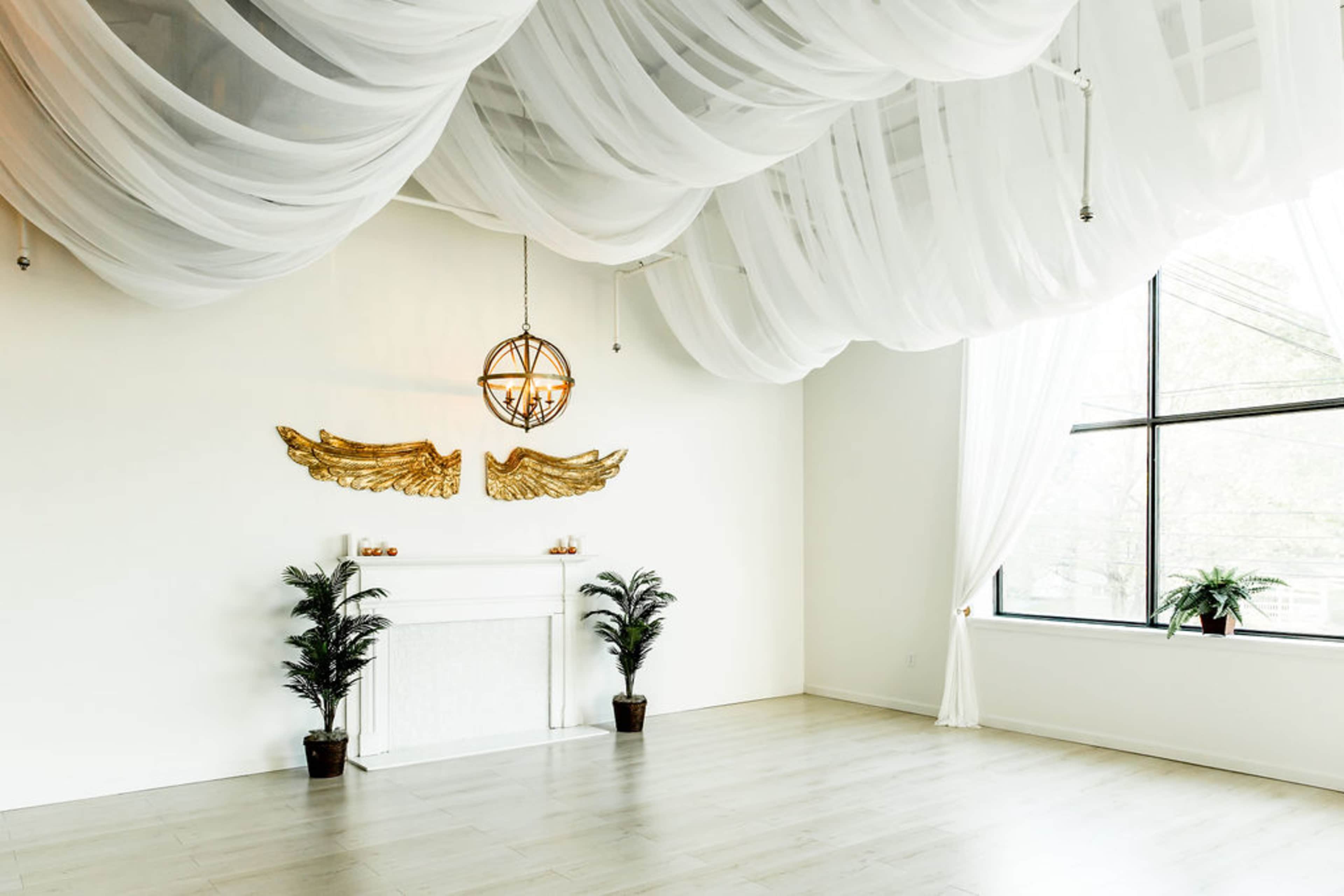 A spacious room with a white draped ceiling, a decorative fireplace, golden wing sculptures on the wall, and large windows allowing natural light to enter.