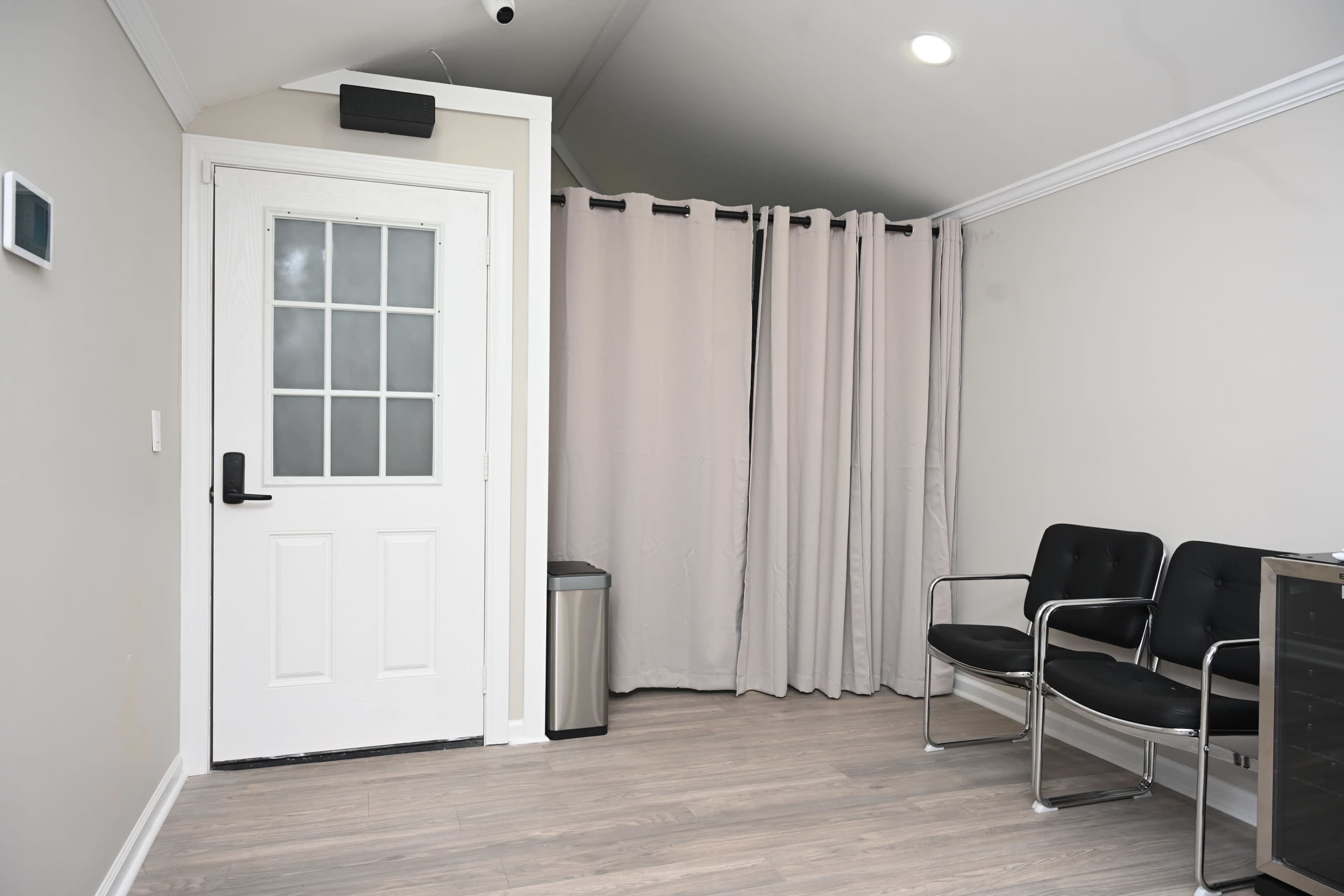 The image shows a small waiting area with a door, a trash can, two black chairs, and a curtain separating an adjacent space.