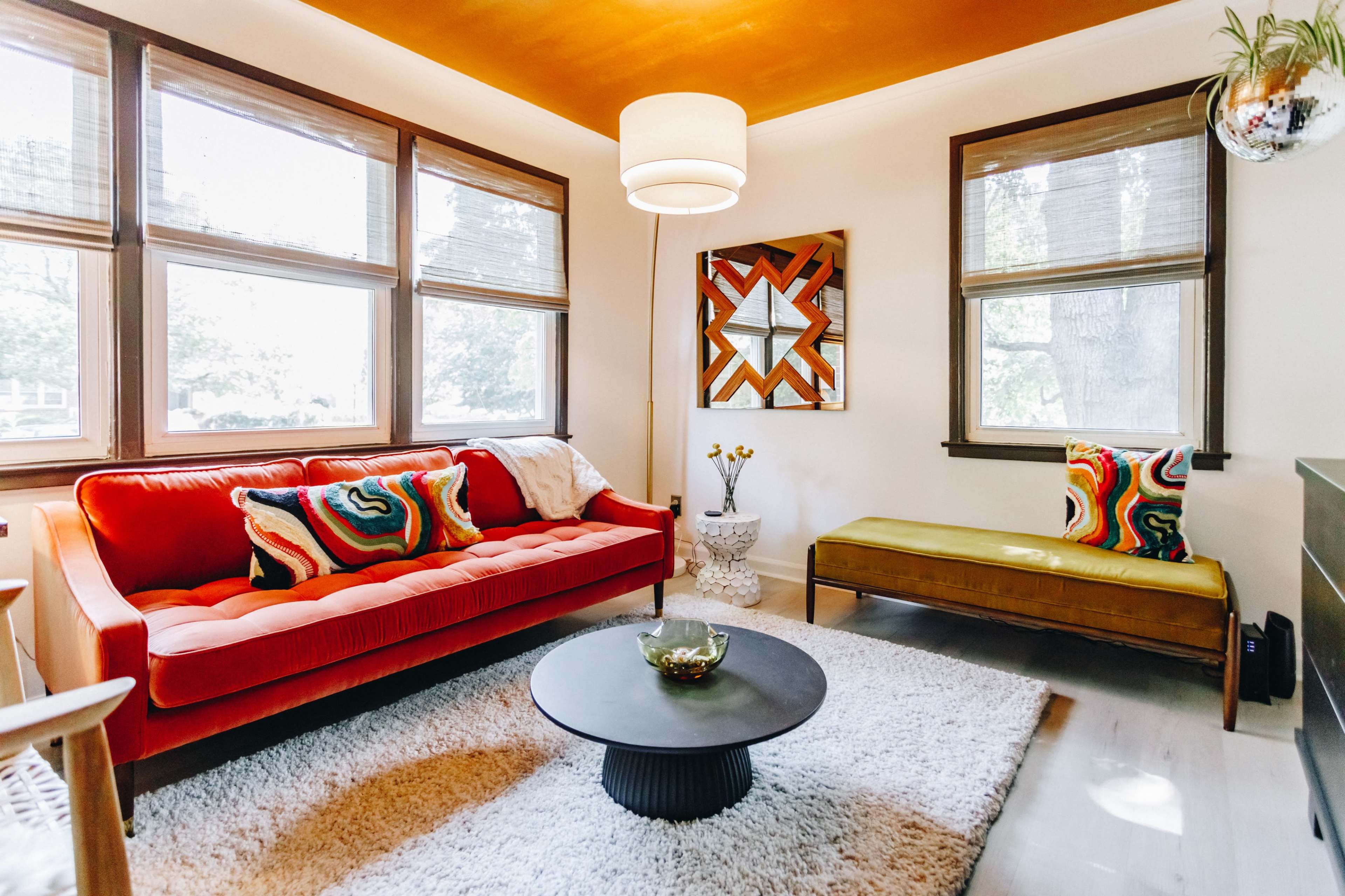A living room with a red velvet sofa, an olive green bench, a round black coffee table, and large windows allowing natural light to enter.