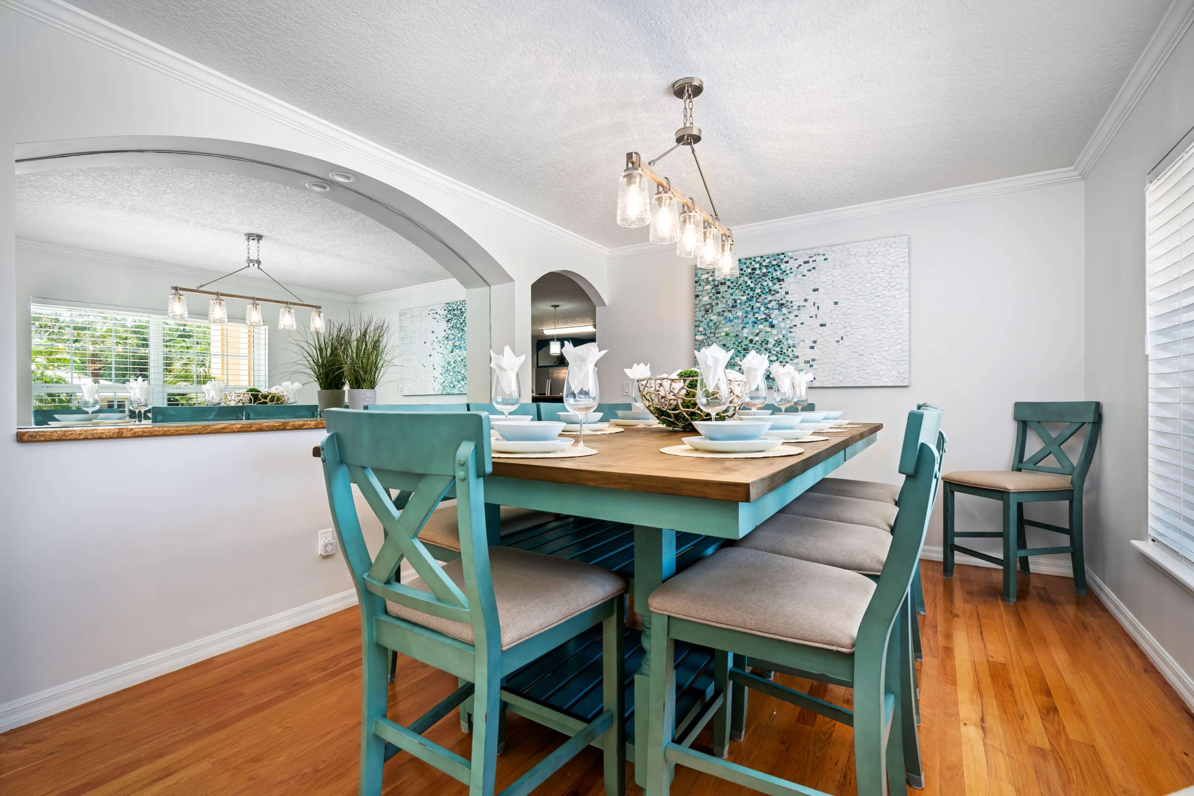 A dining area features a wooden table surrounded by teal chairs, with place settings and a modern light fixture above.