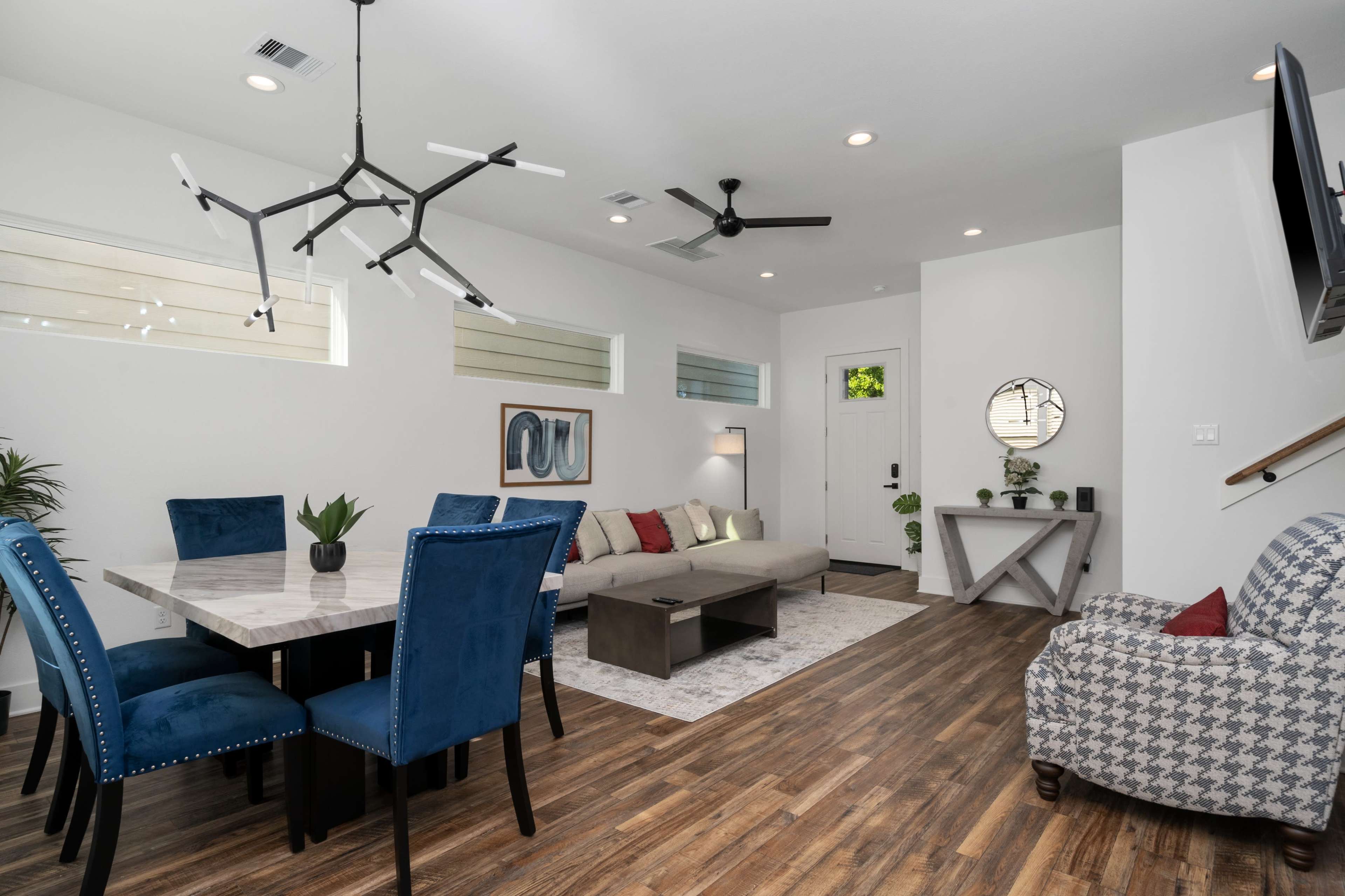 A modern living and dining area features a table with blue chairs, a gray sofa, and a wall-mounted TV.