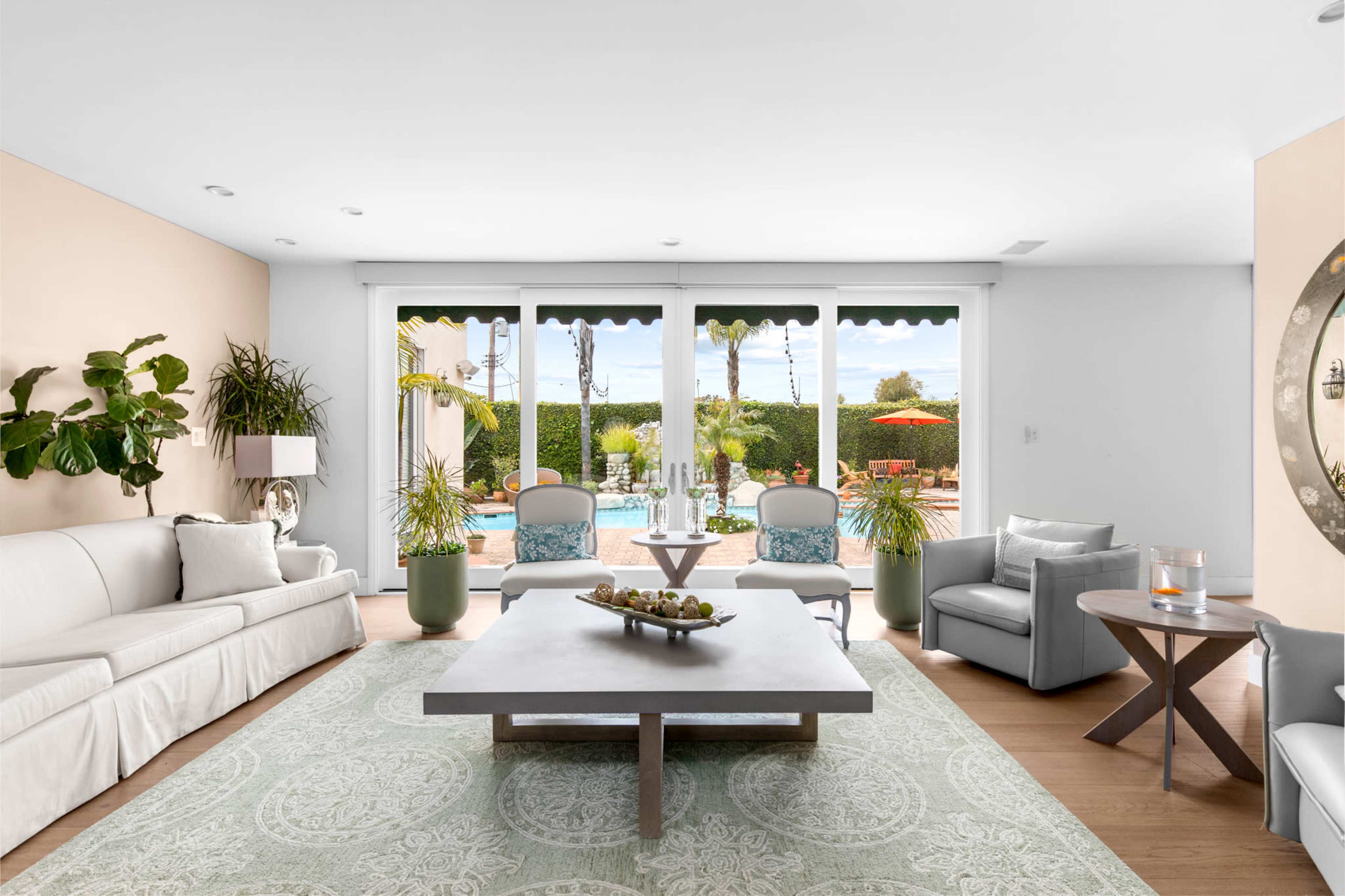 The image shows a bright living room with large windows overlooking a pool area, furnished with a light sofa, gray chairs, and a central coffee table on a decorative rug.
