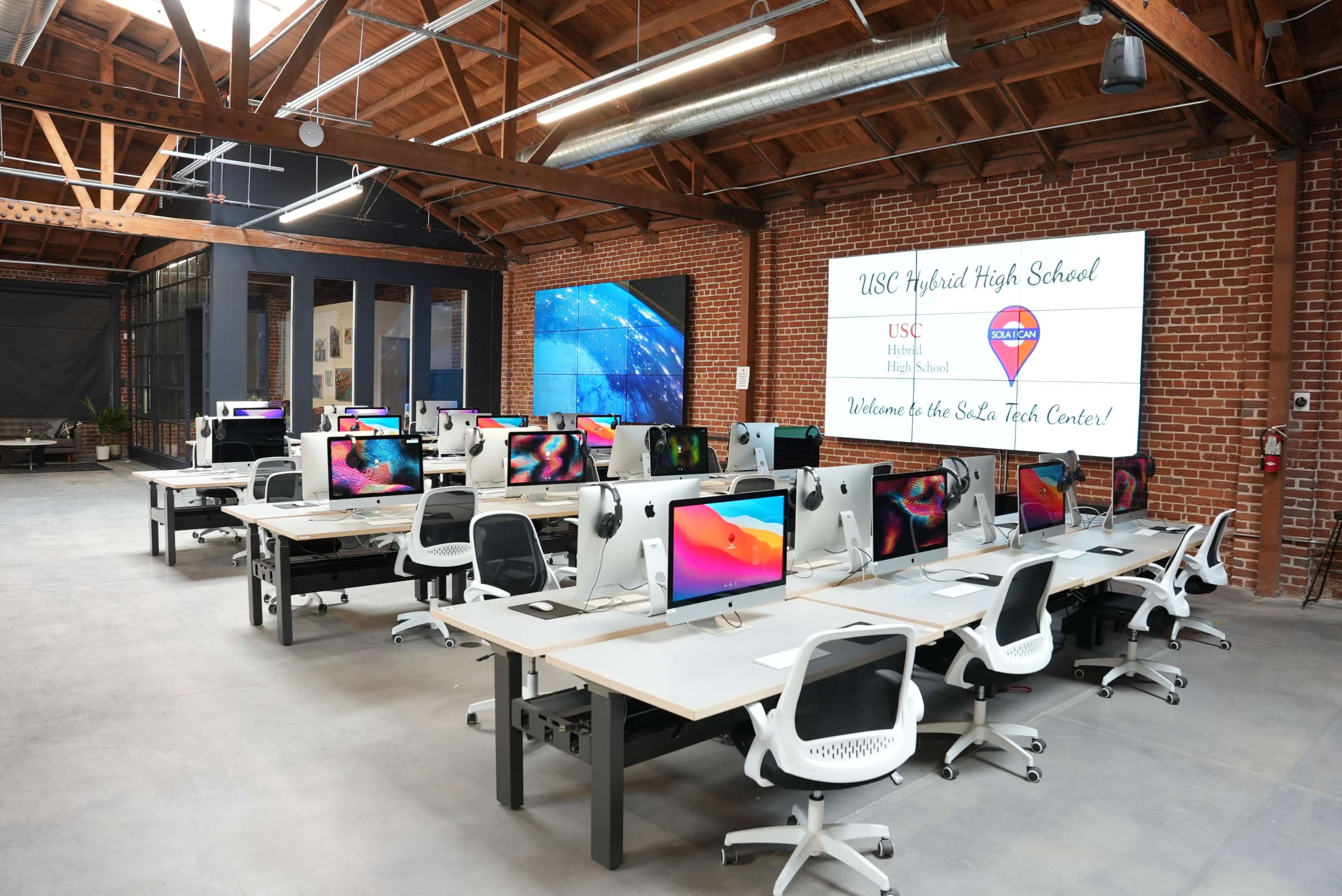 A modern tech center at USC Hybrid High School features multiple computer stations arranged with iMacs on sleek desks under a wooden ceiling.