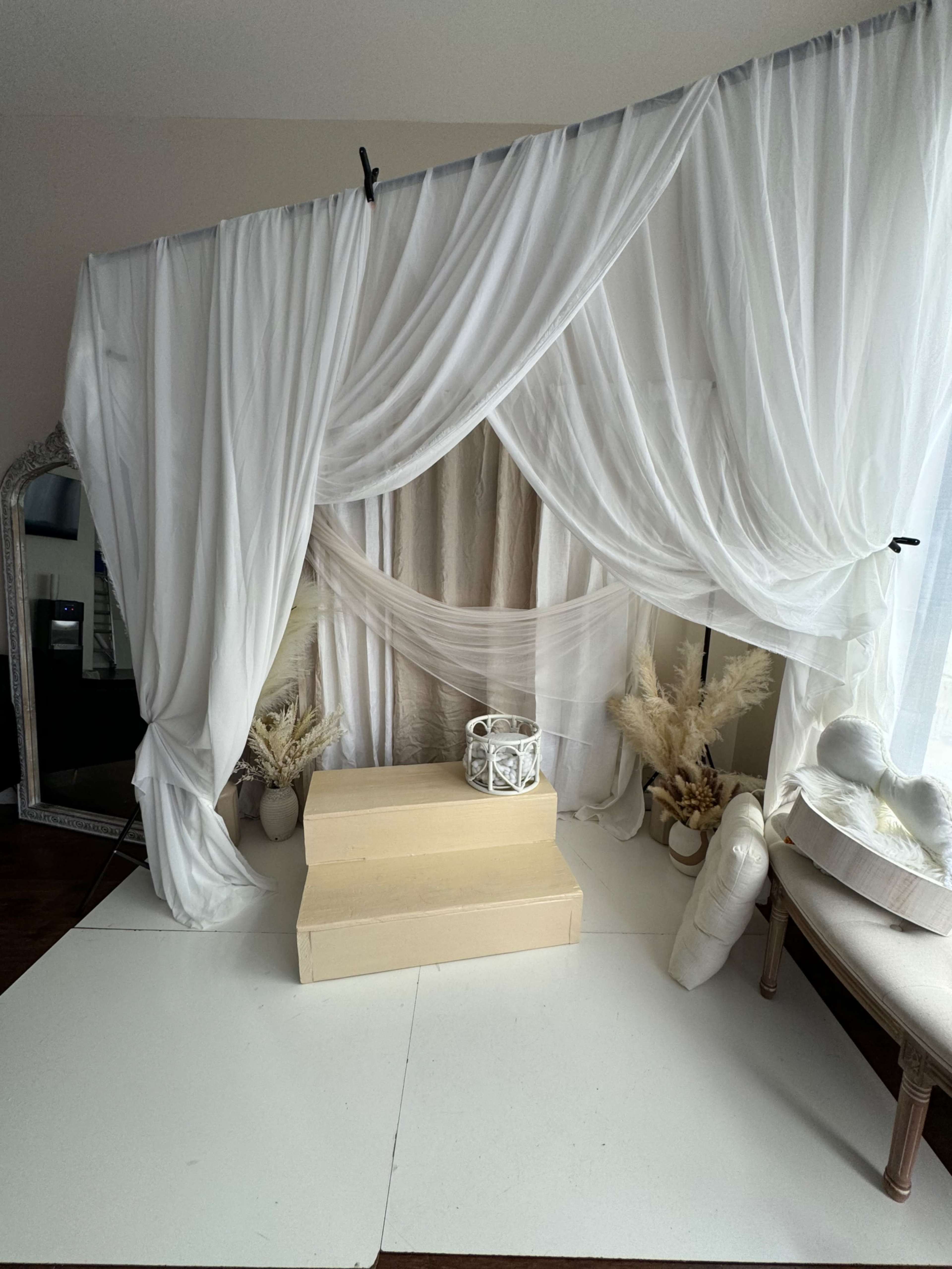 A staged area with white drapes, a light-colored platform, and decorative elements including a mirror and potted plants.