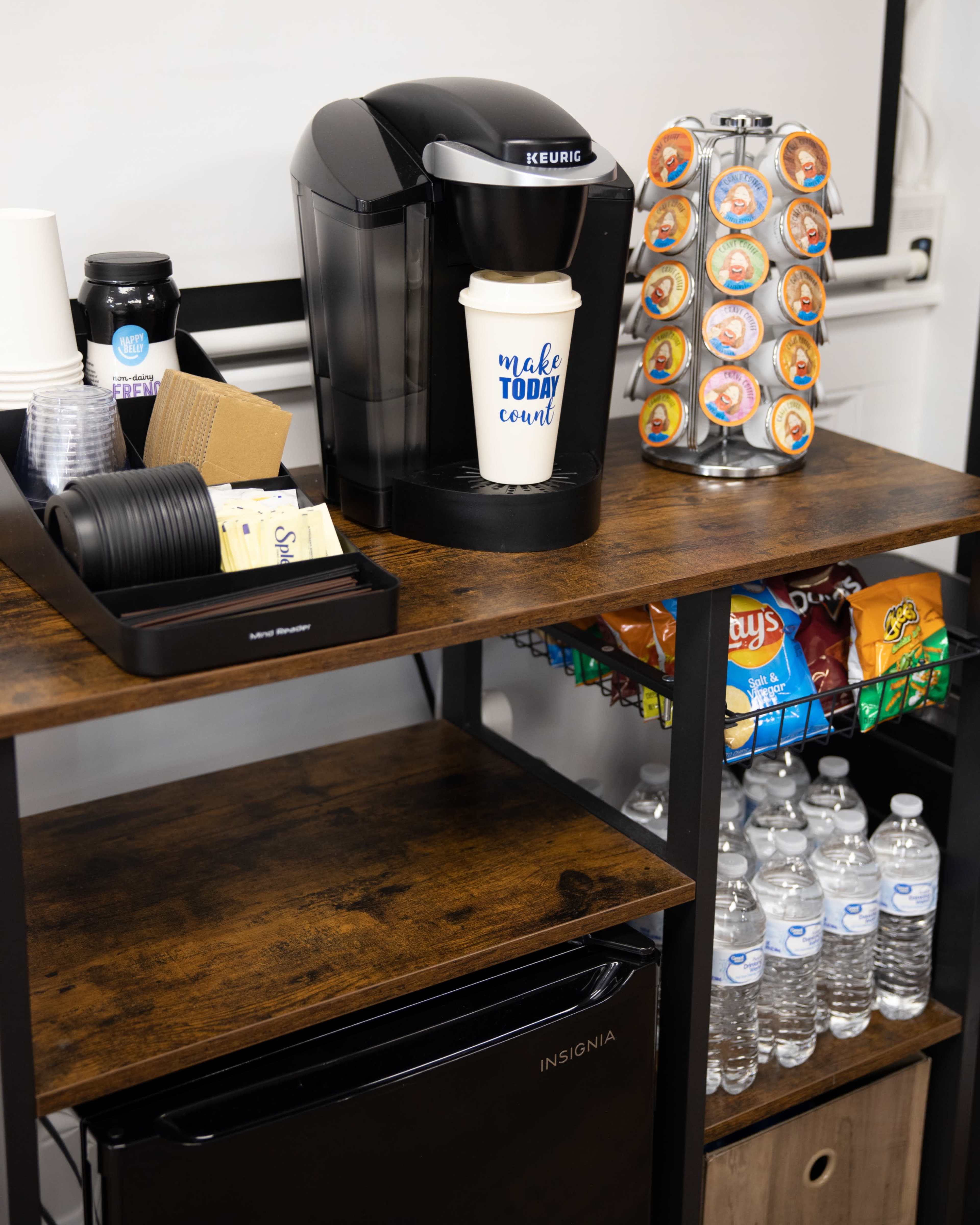 A coffee station featuring a Keurig machine, coffee cups, various creamers, snacks, and bottles of water on wooden shelves.