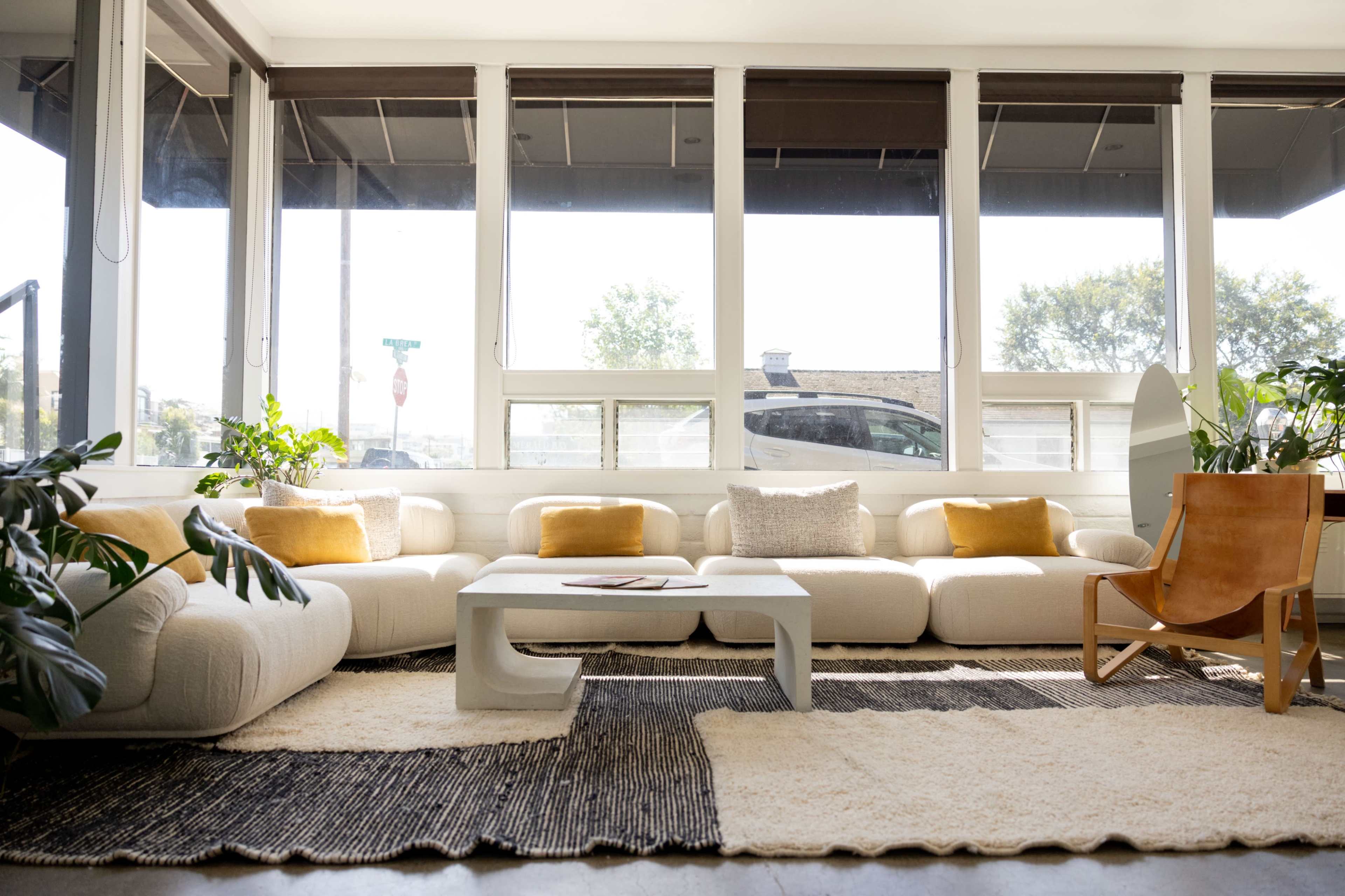 The image shows a bright, modern living room with a large, white sectional sofa, a coffee table, and various plants placed around the space.
