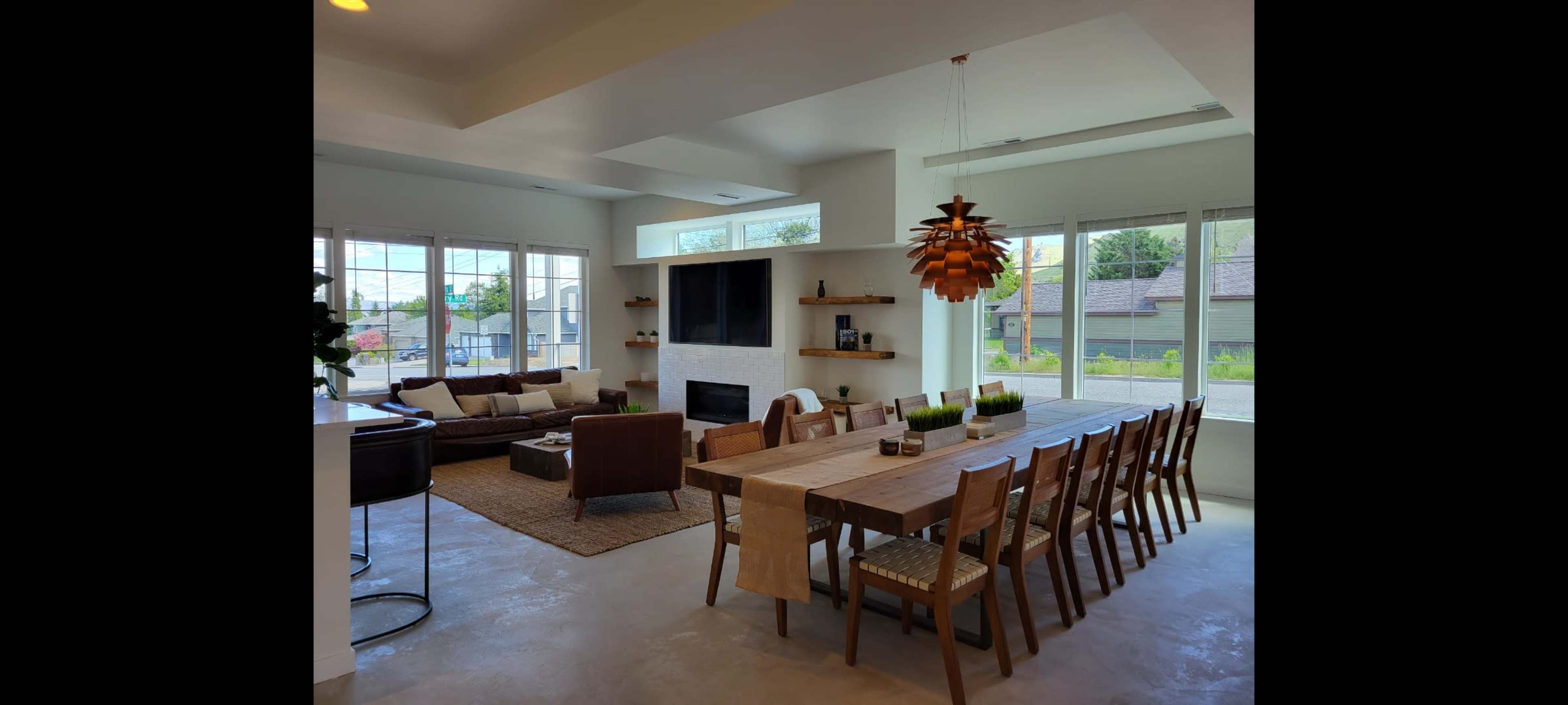 A modern dining area with a long wooden table surrounded by chairs, adjacent to a spacious living room featuring a large sofa and a wall-mounted television.