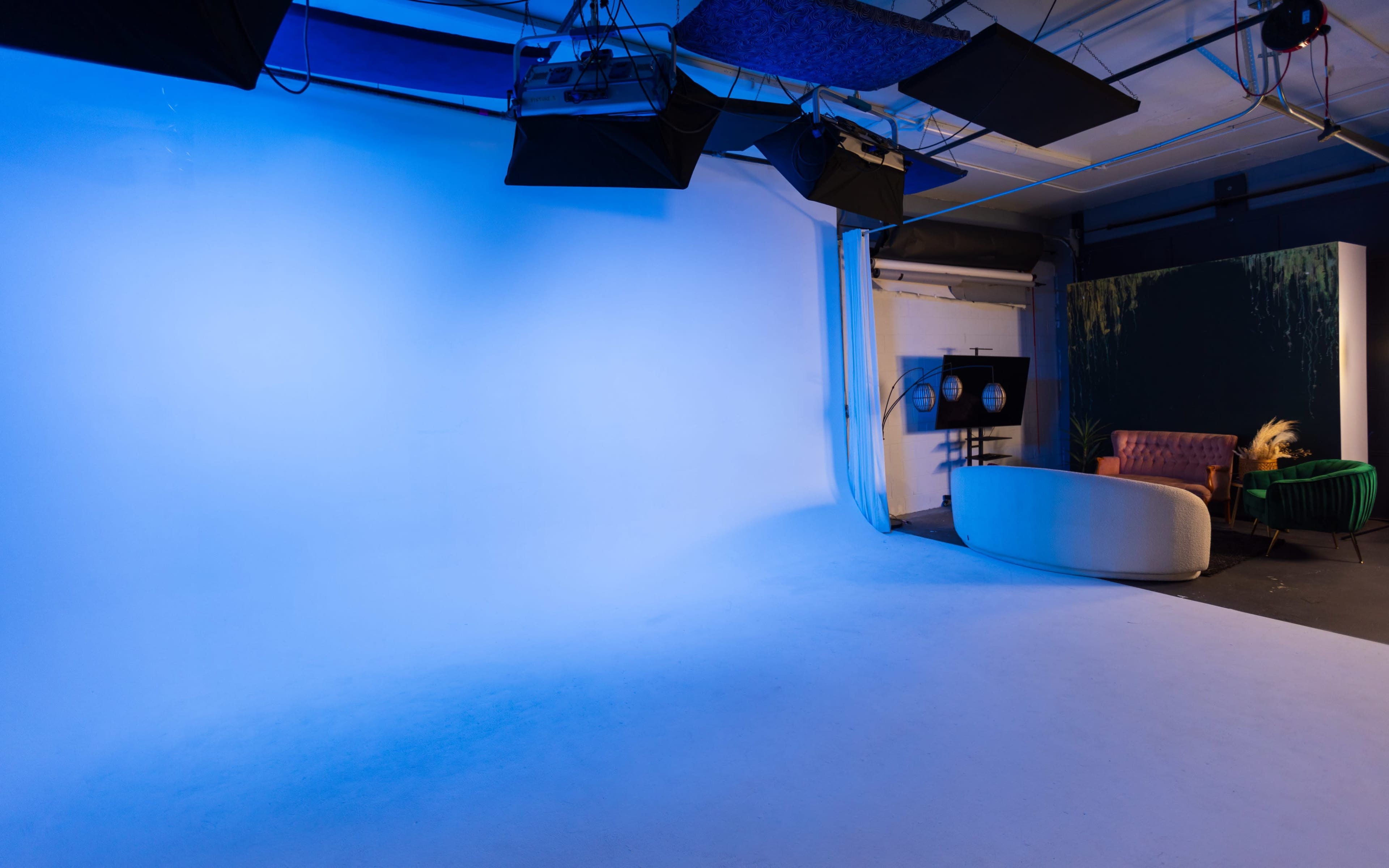 A photo studio with a blue backdrop, lighting equipment, and a minimalistic set featuring a pink sofa and a green armchair.