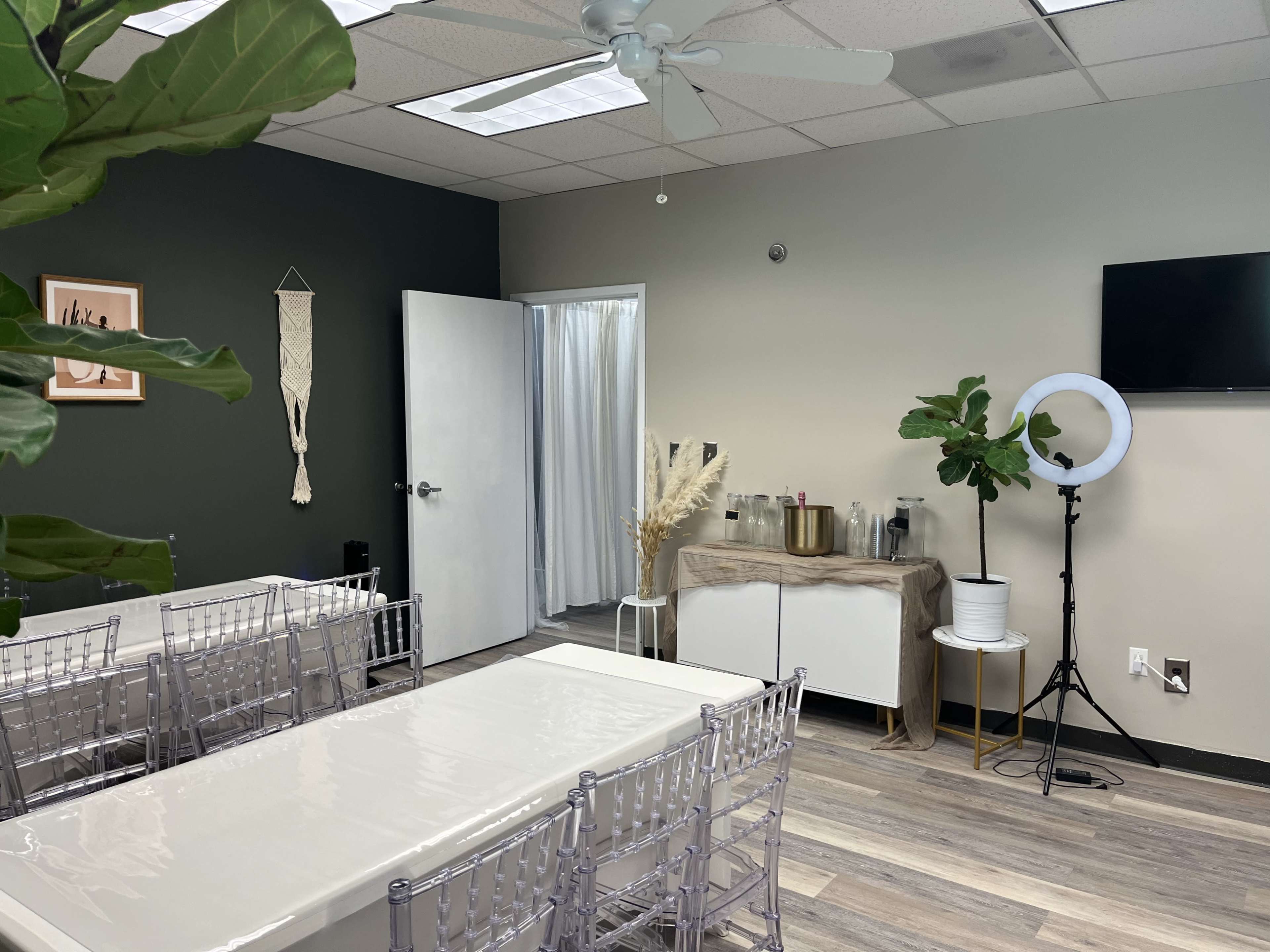 A neatly arranged meeting room with a large table, transparent chairs, a plant, a wall-mounted TV, and a bar area featuring drinks and decorative elements.