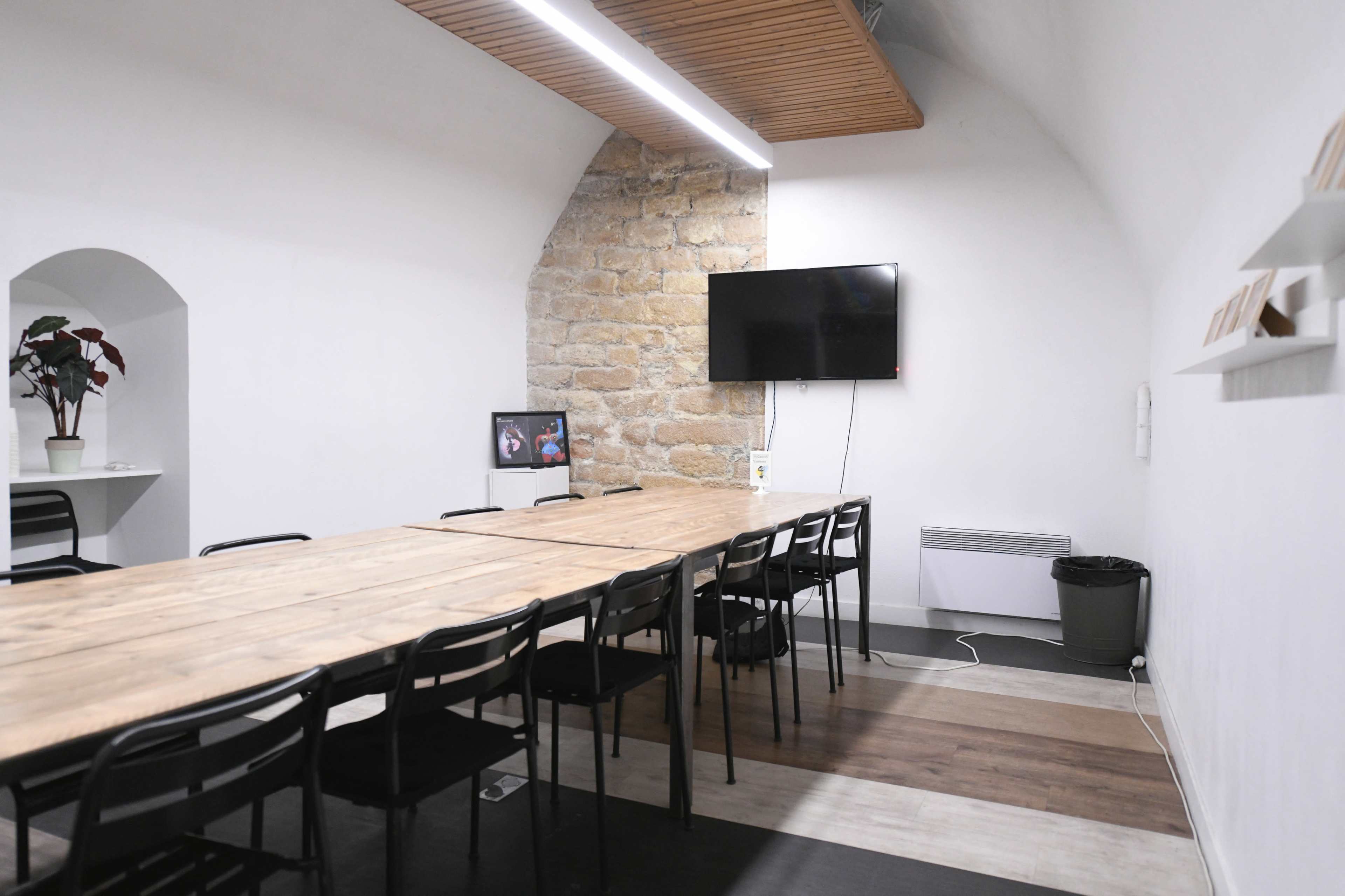 A modern meeting room features a long wooden table surrounded by black chairs, with a stone wall and a mounted television on one side.