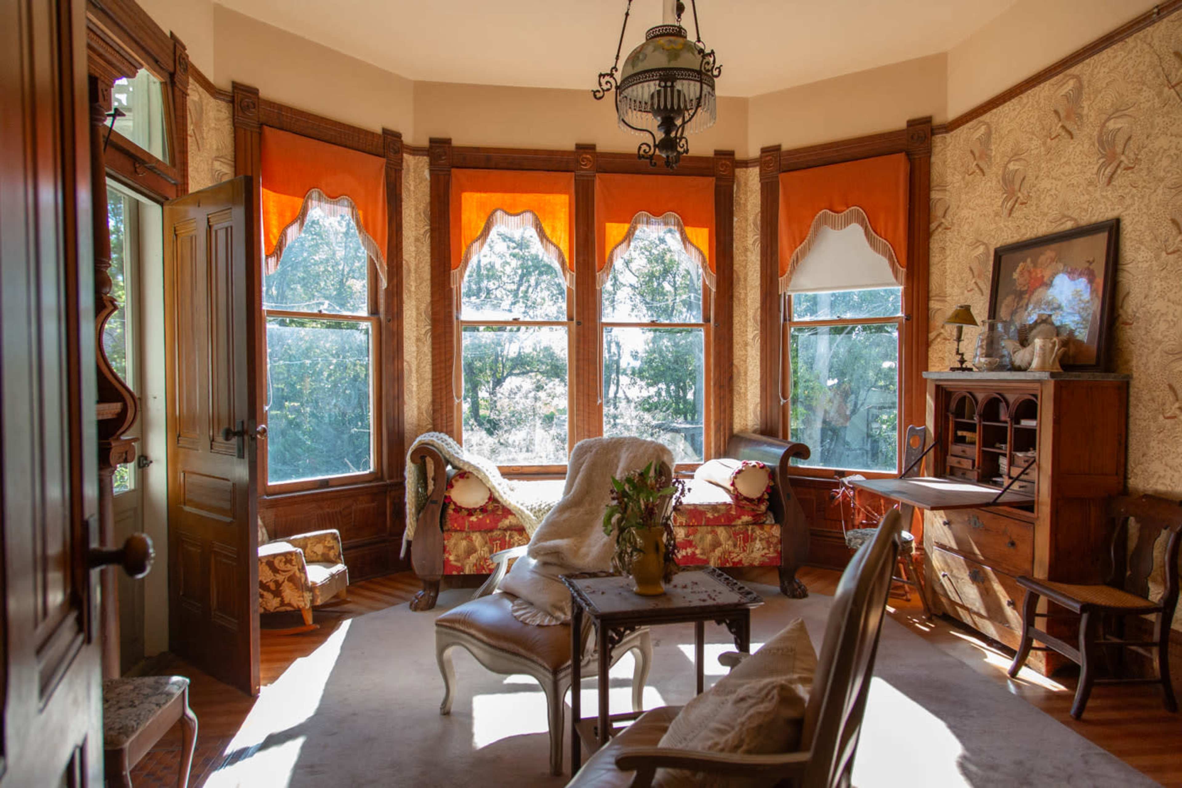 The image shows a well-lit room with large windows dressed in orange curtains, featuring vintage furniture and a decorative table.