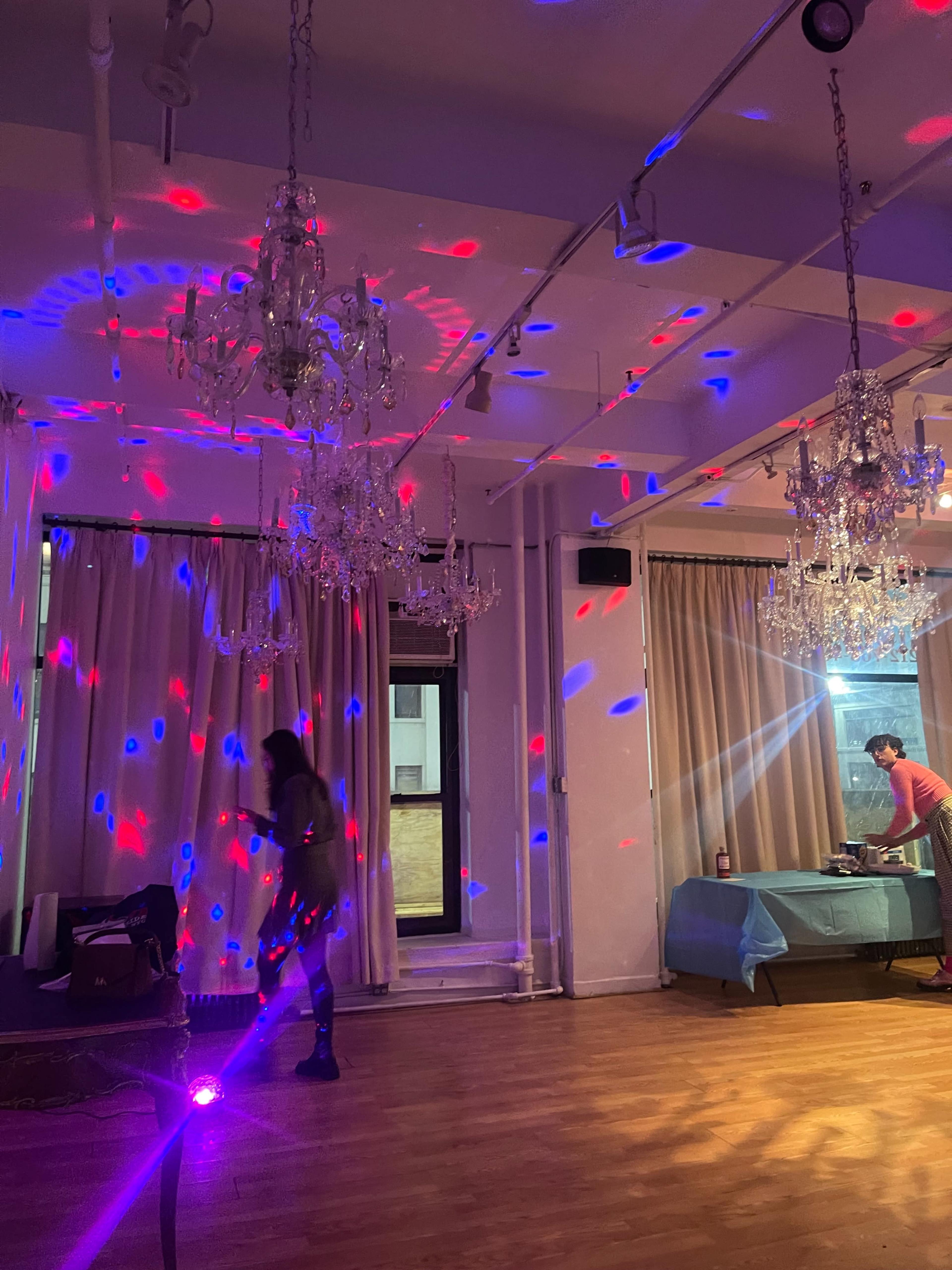 A dance floor with colorful lights reflecting off the walls and two chandeliers, where a person is dancing and another is setting up equipment.