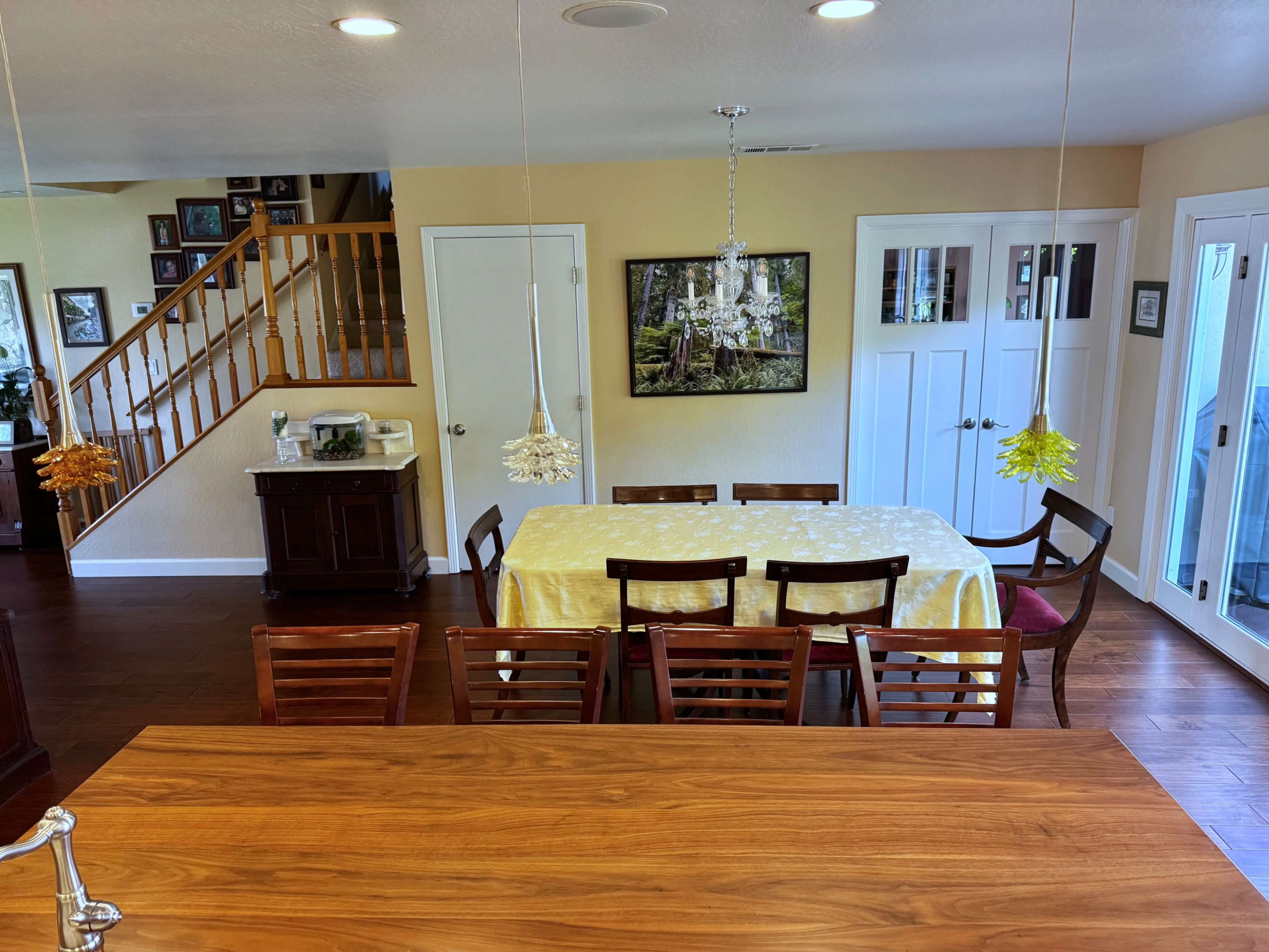 The image shows a dining area with a large table set for meals, surrounded by wooden chairs, and featuring a staircase and decorative wall framing in the background.