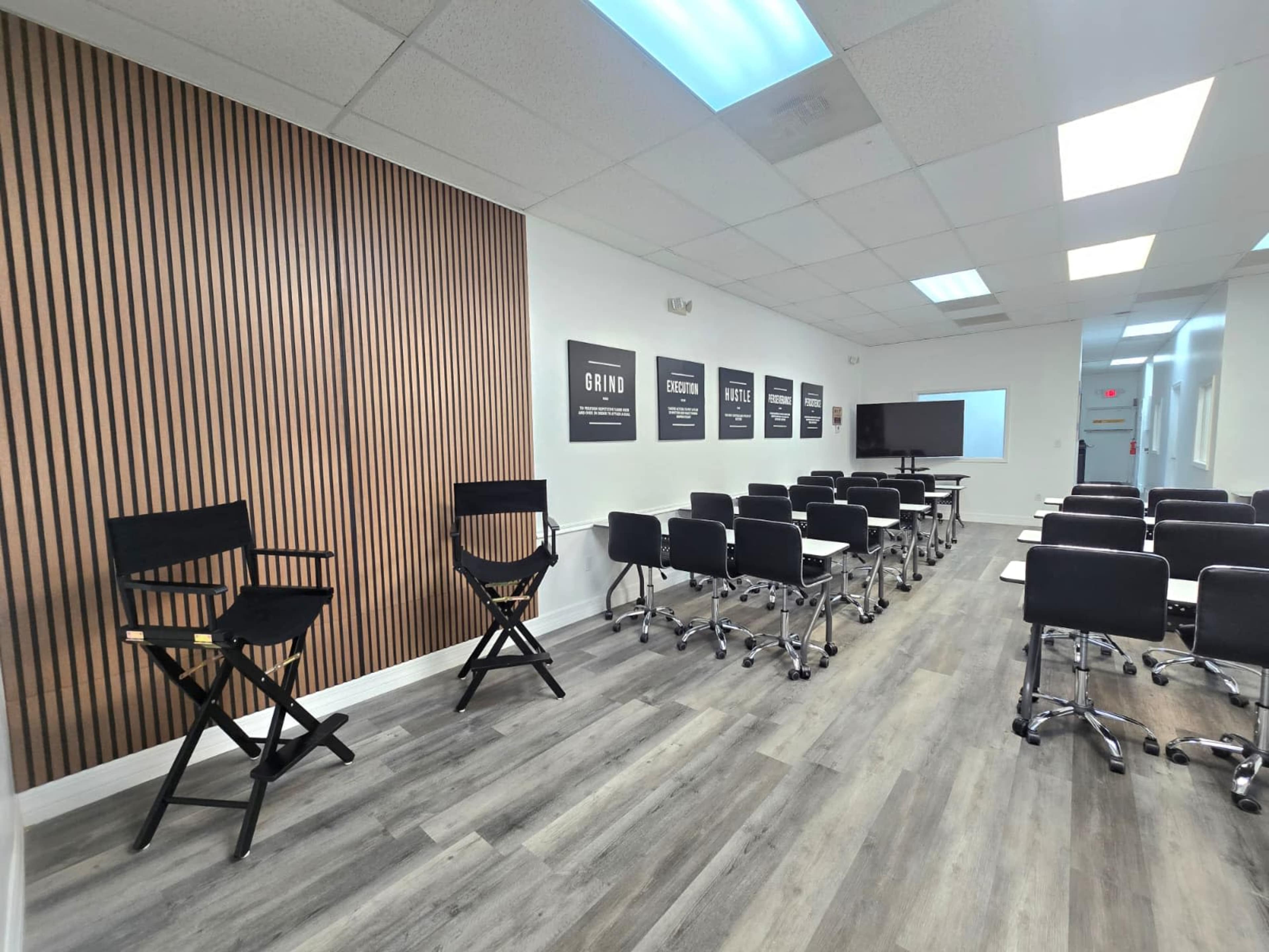 The image shows a modern classroom with black rolling chairs arranged in rows, a wooden accent wall, and a screen at the front.