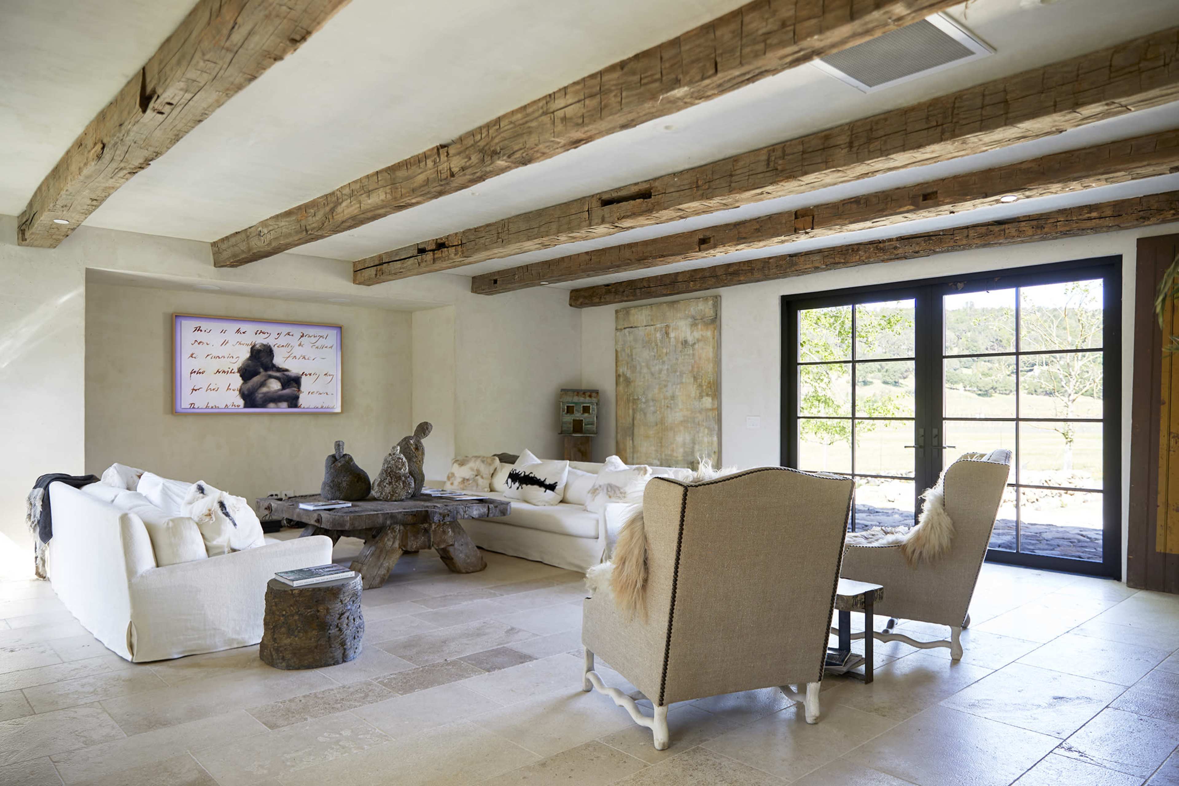 A spacious living room features two white sofas, a round wooden coffee table, and a pair of armchairs, with large glass doors opening to a garden view.