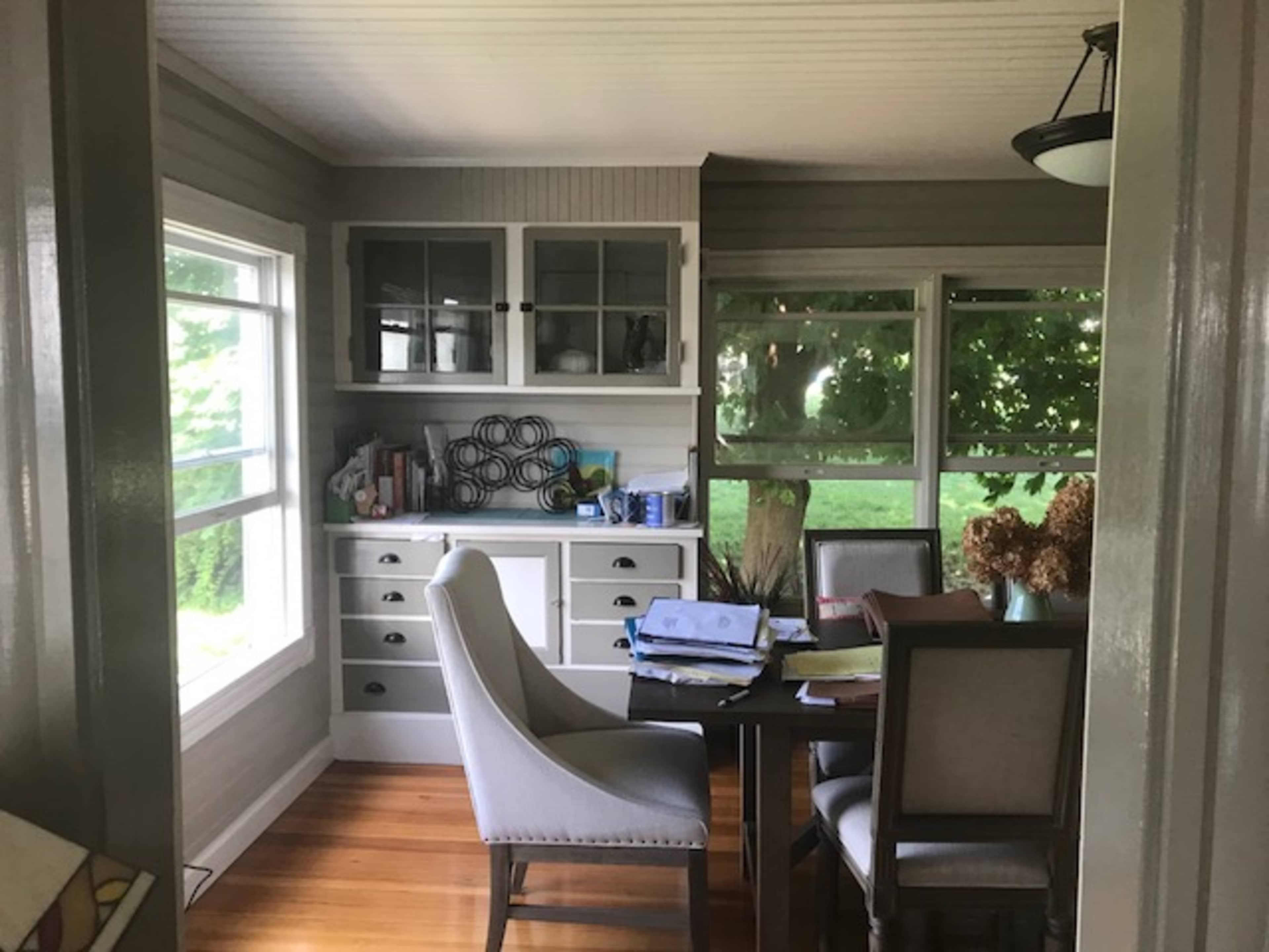 The image shows a neatly organized dining area with a table and chairs, adjacent to a windowed space featuring a cabinet and various items on the counter.