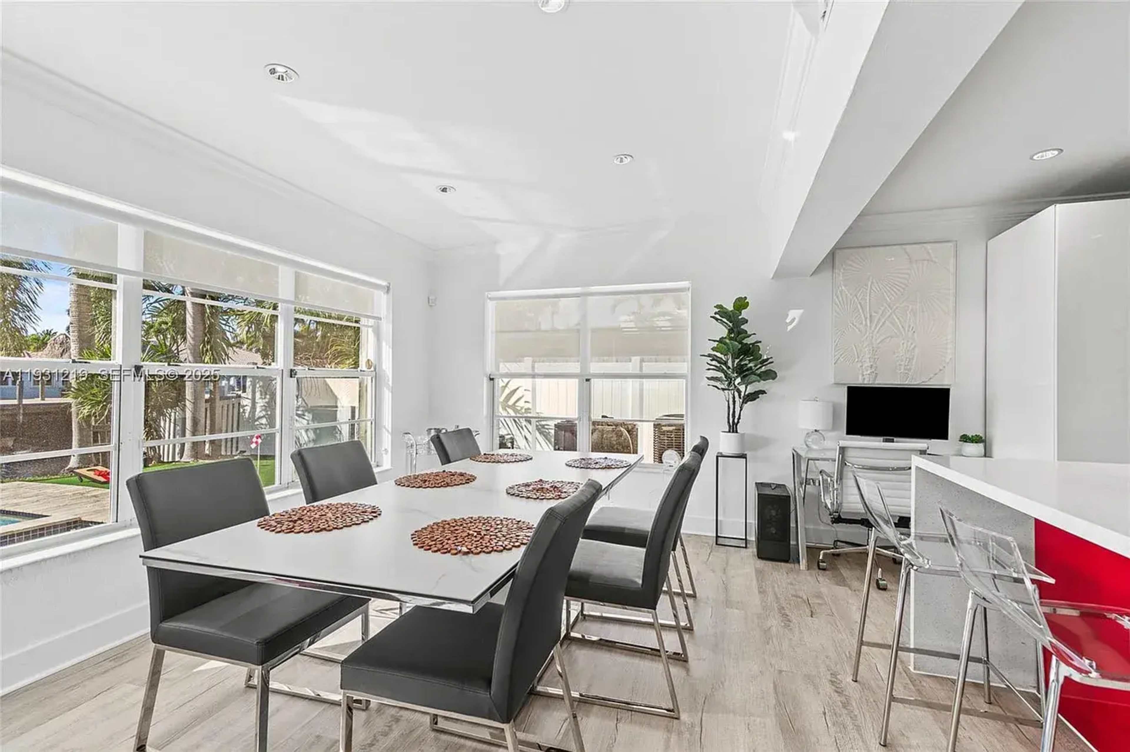 The image shows a modern dining area with a white table set for six, surrounded by sleek metallic chairs and large windows allowing natural light to illuminate the space.