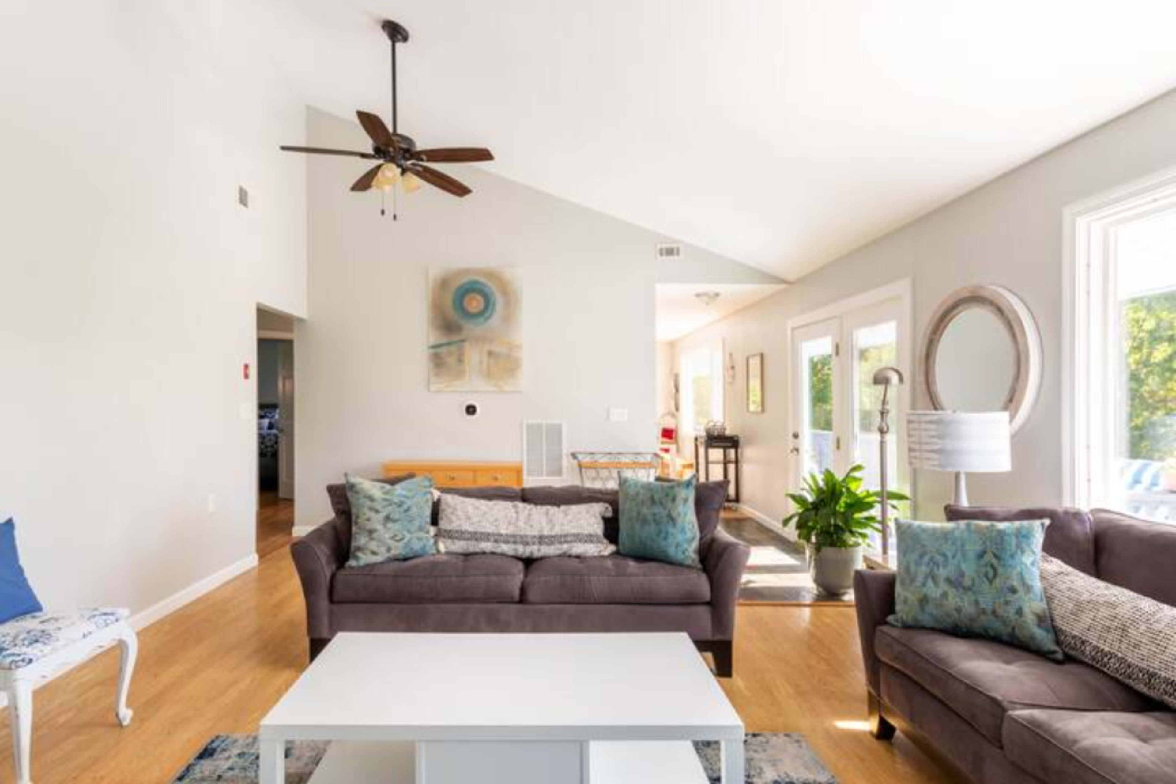 A bright living room features a ceiling fan, two sofas with decorative pillows, a coffee table, and large windows that provide natural light.