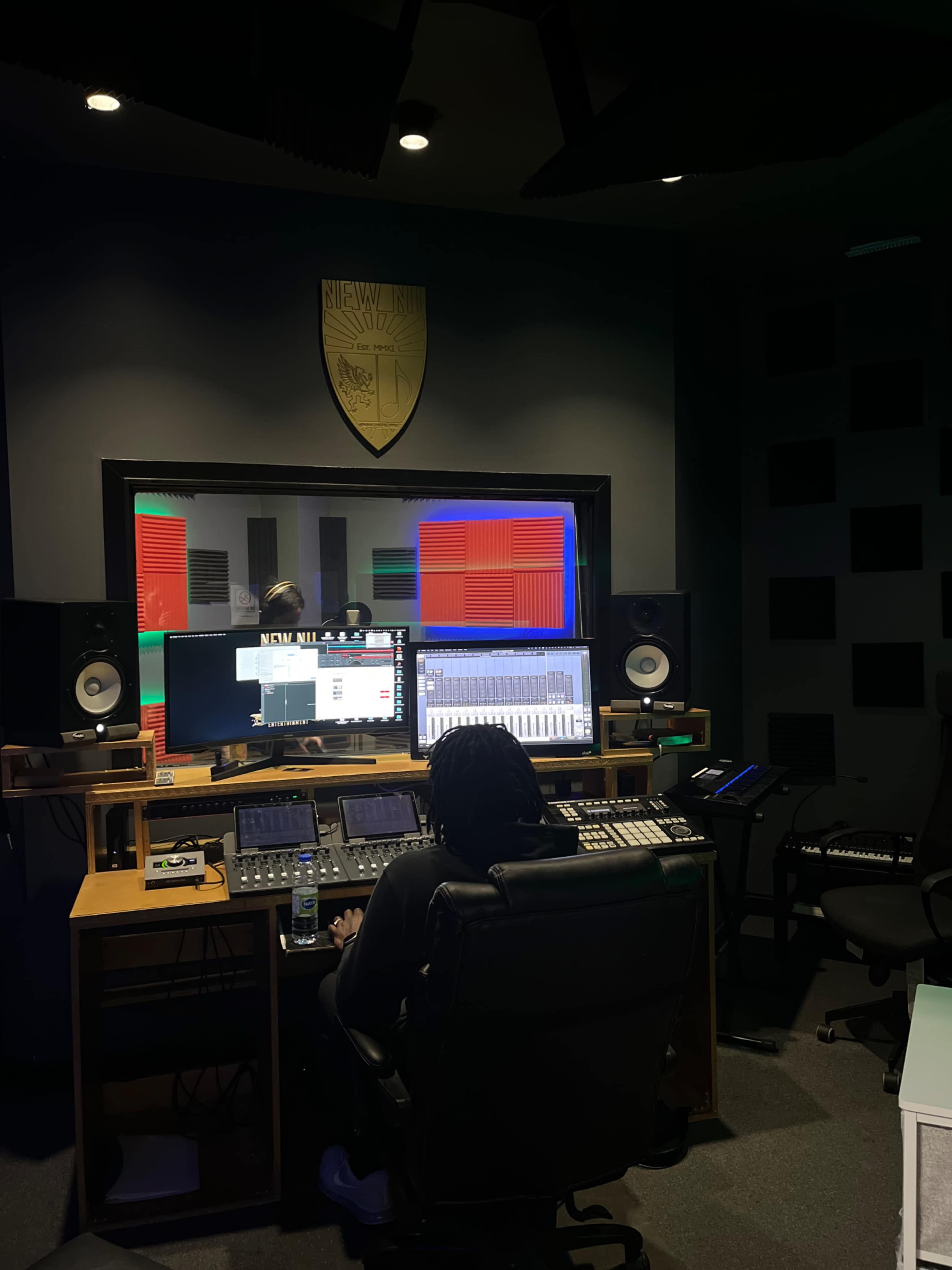 A person is working at a music production console in a studio with computer screens and audio equipment.